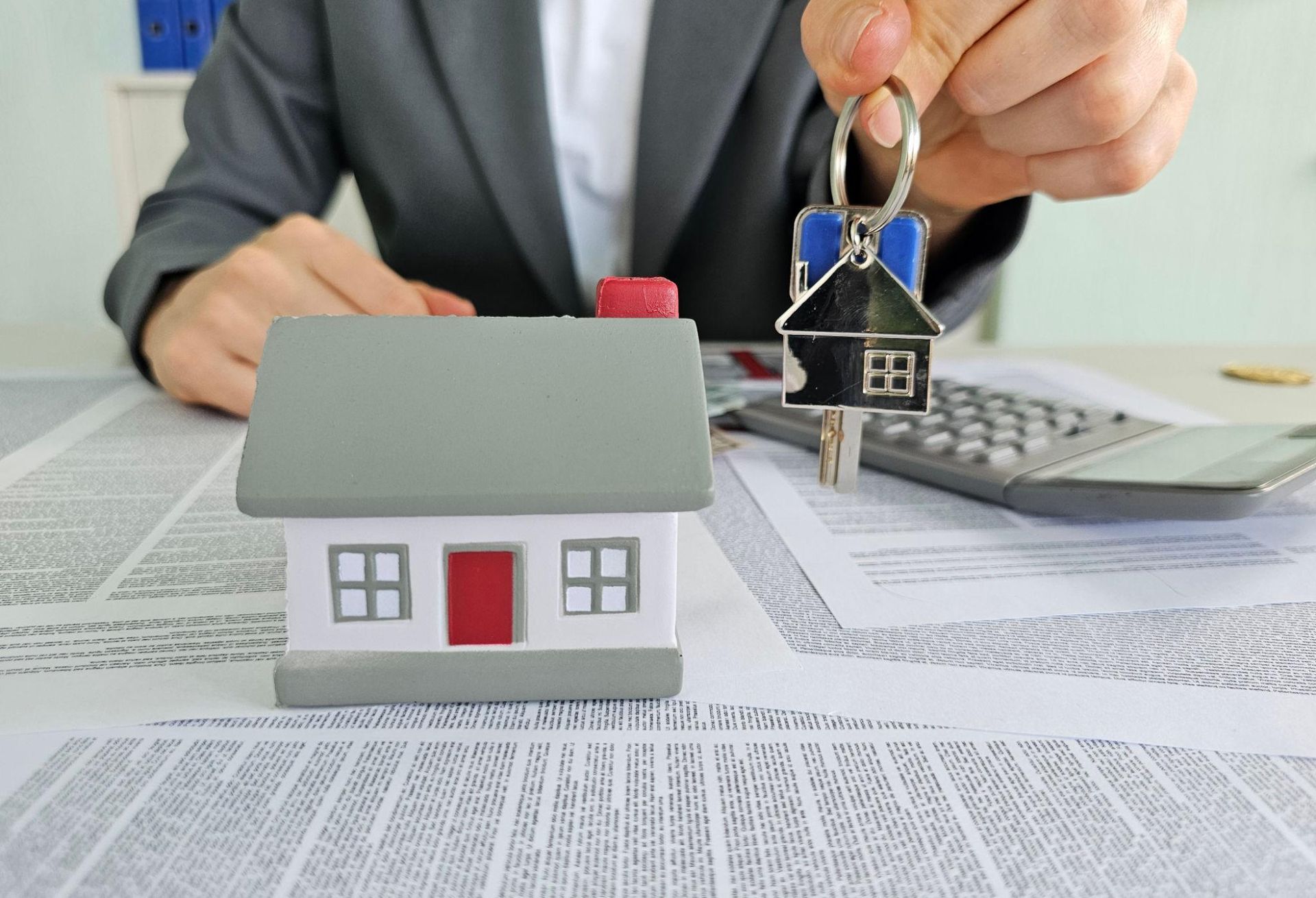 Hand holding house keys over a model house, with calculator and paperwork in the background