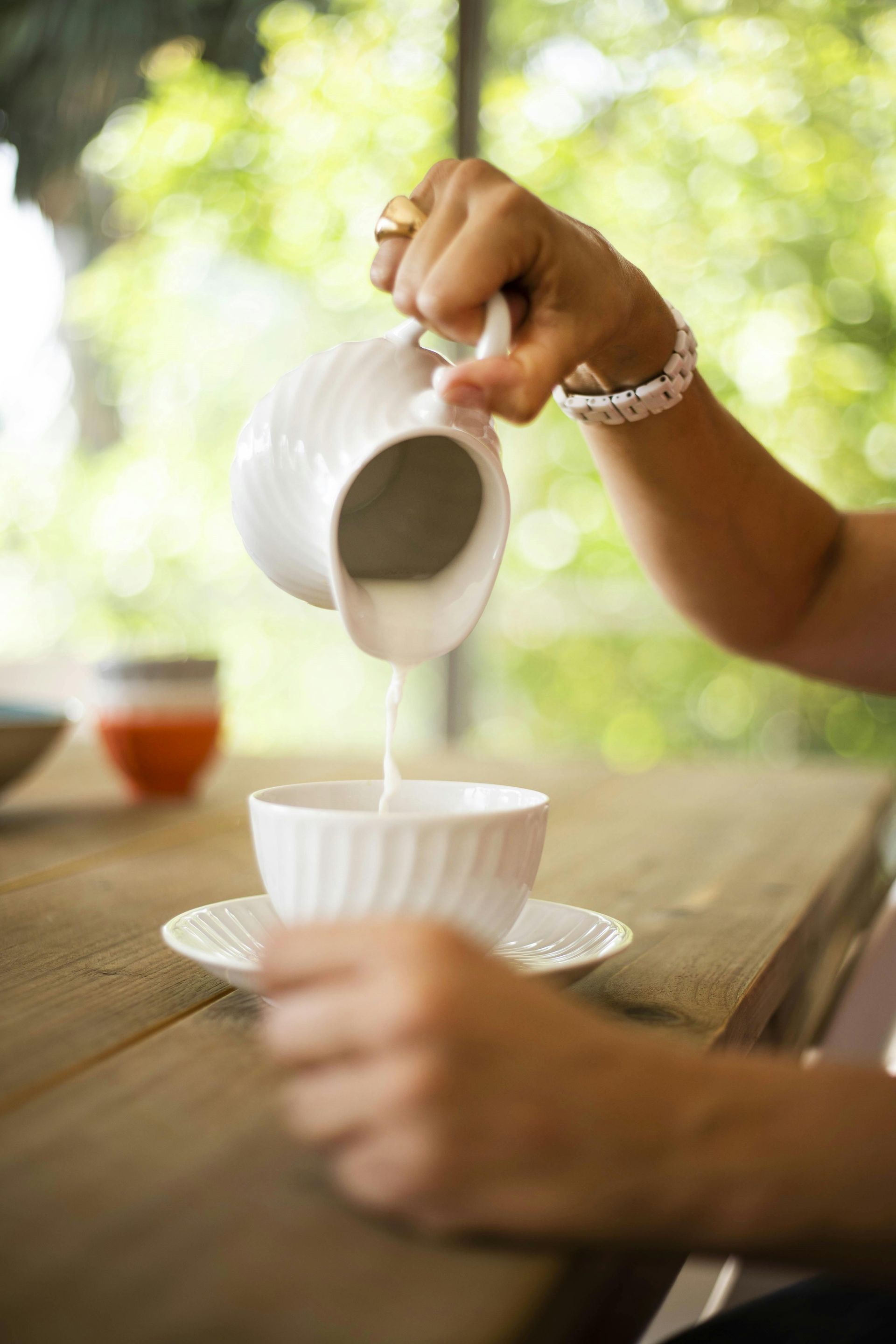 Hand pouring milk from a white pitcher into a cup on a wooden table outdoors