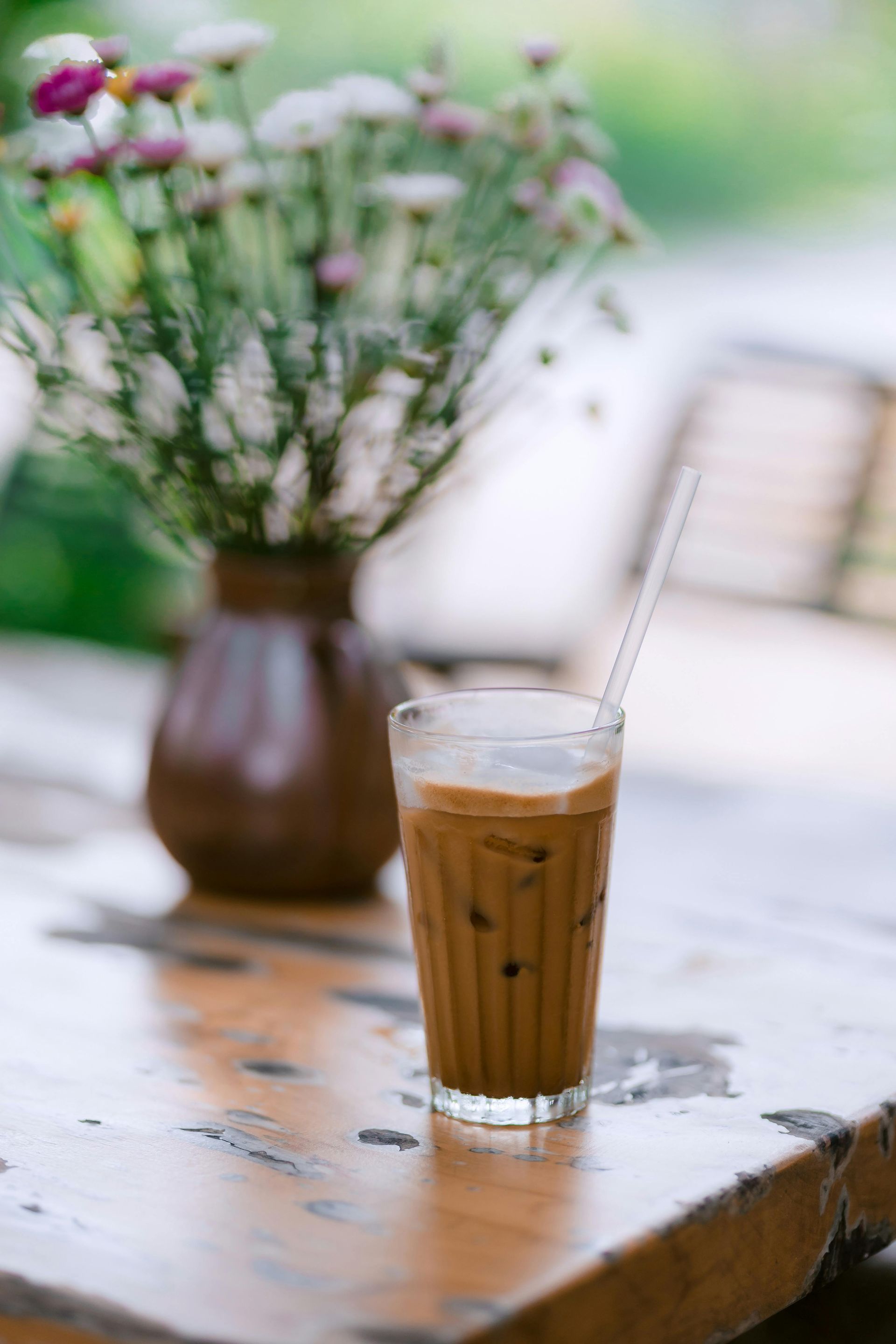 Iced coffee in a glass on a rustic wooden table with a blurred vase of flowers in the background