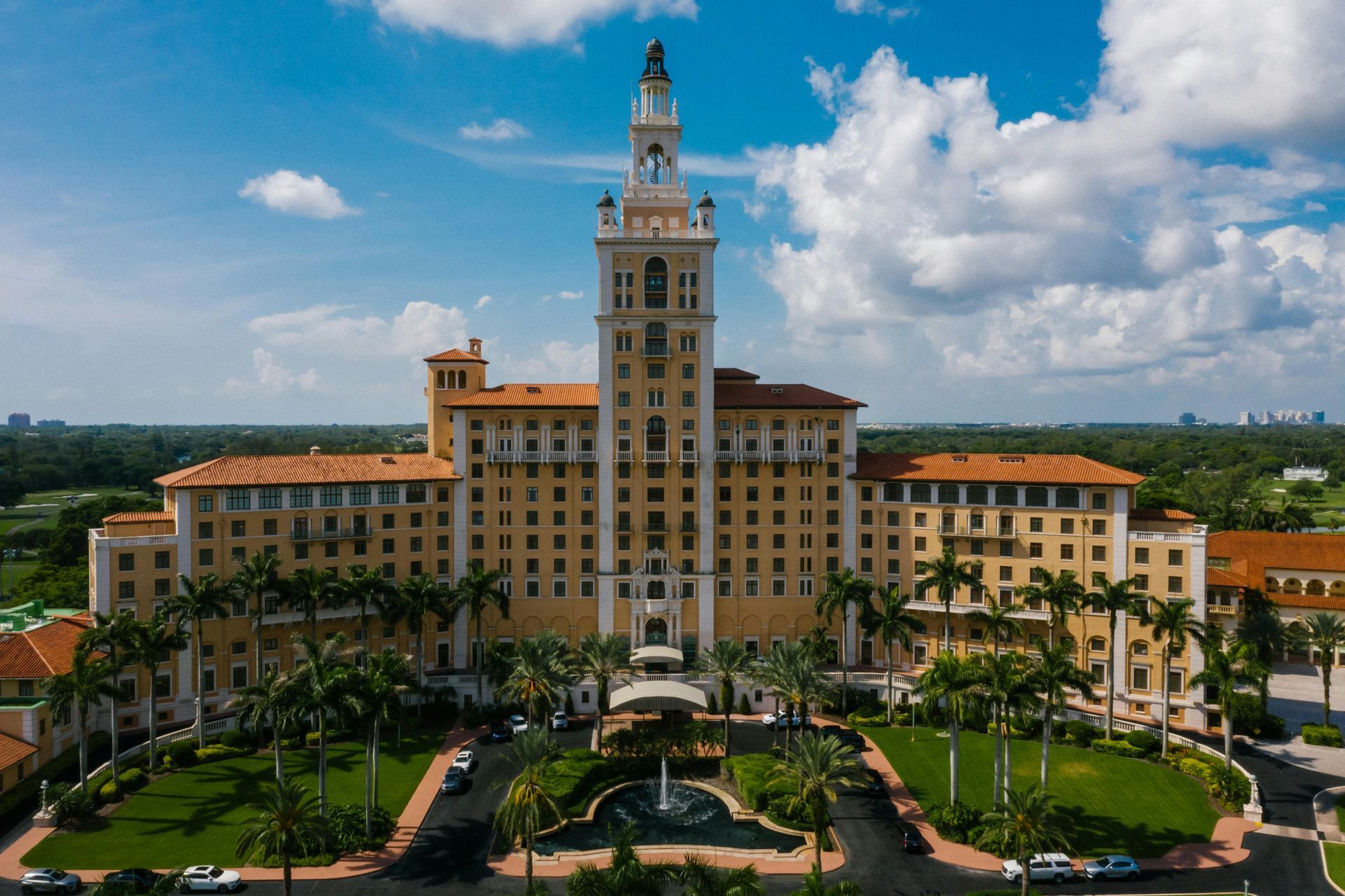 Large historic hotel with a central tower, red-tiled roofs, and palm-lined gardens under a blue sky.