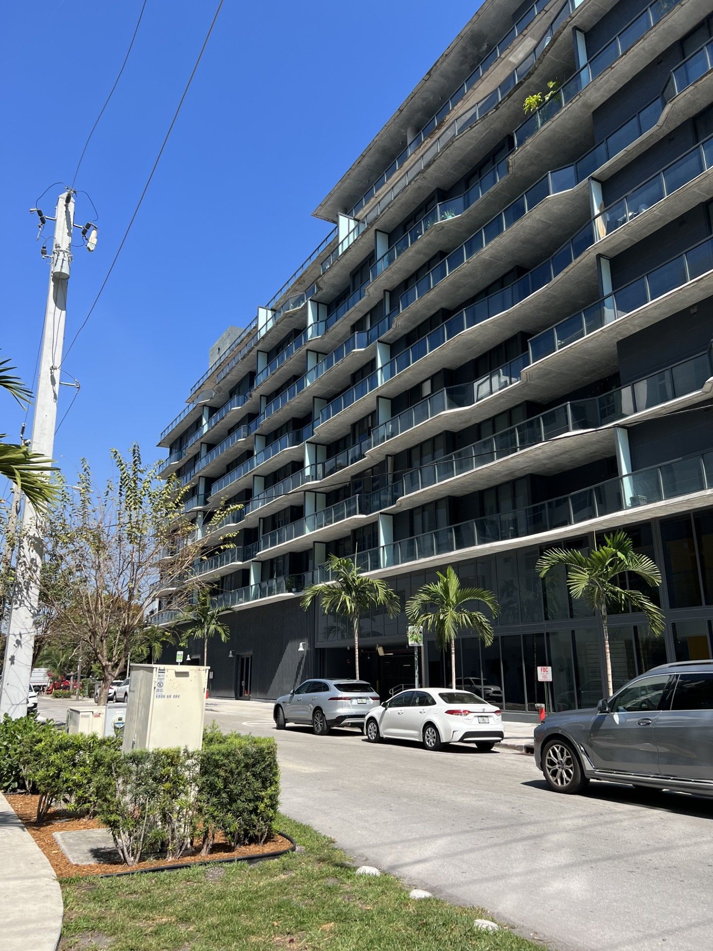 Modern apartment building with balconies, parked cars, and trees along a sunny street