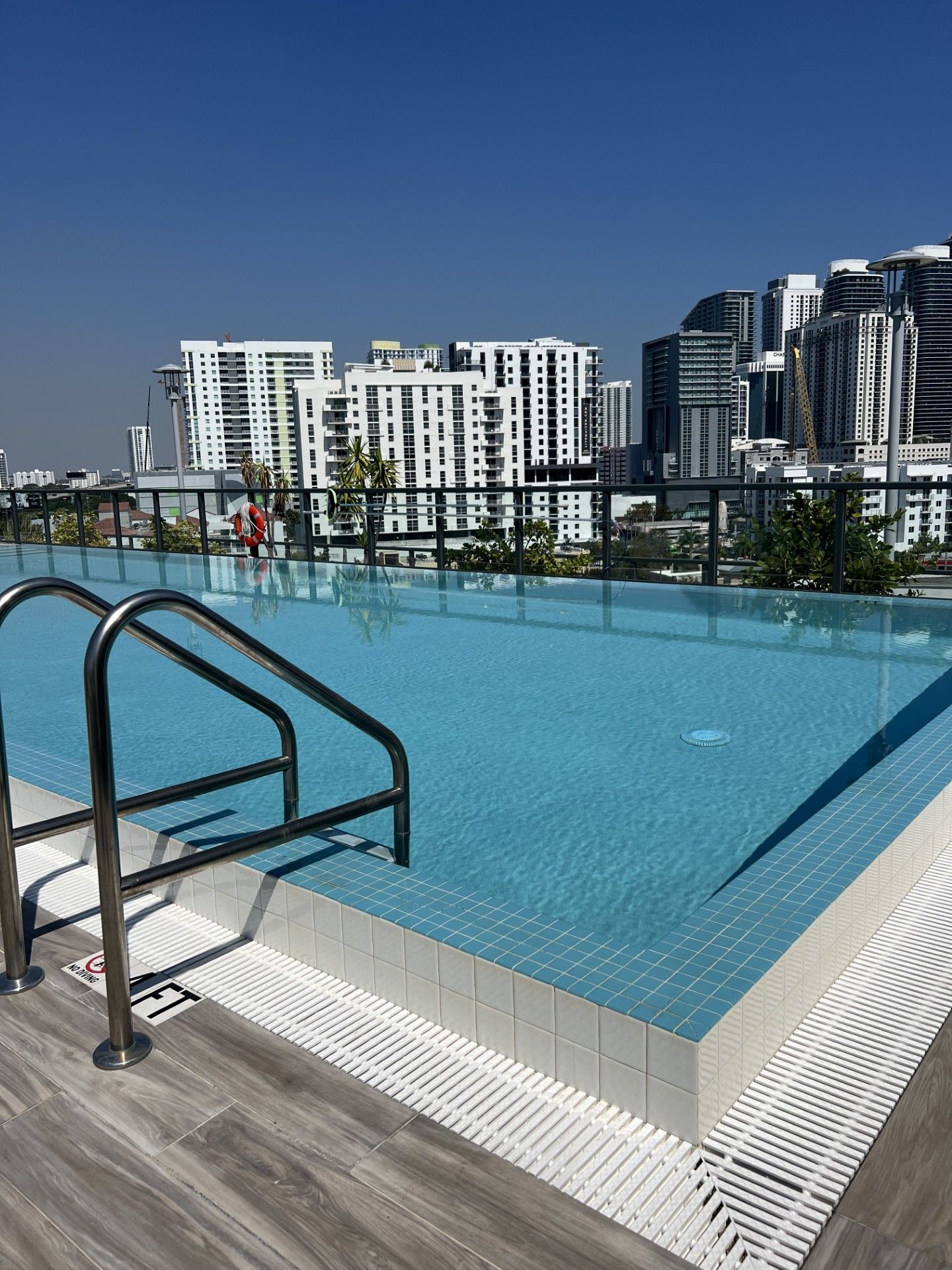 Rooftop infinity pool with turquoise water, metal handrail, and city skyline under a clear blue sky
