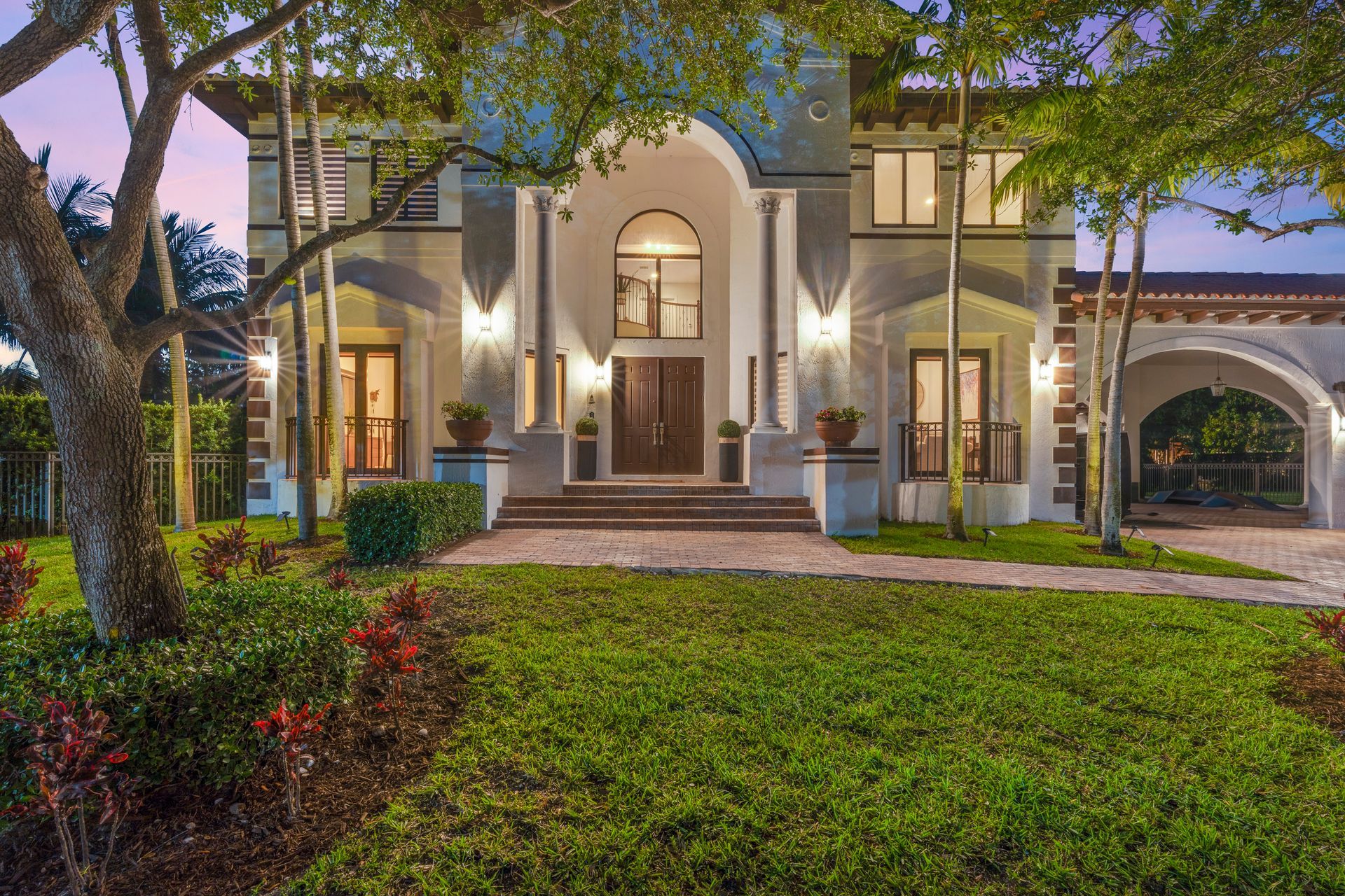 Elegant two-story house with lit entryway, arched windows, and a manicured front lawn at dusk