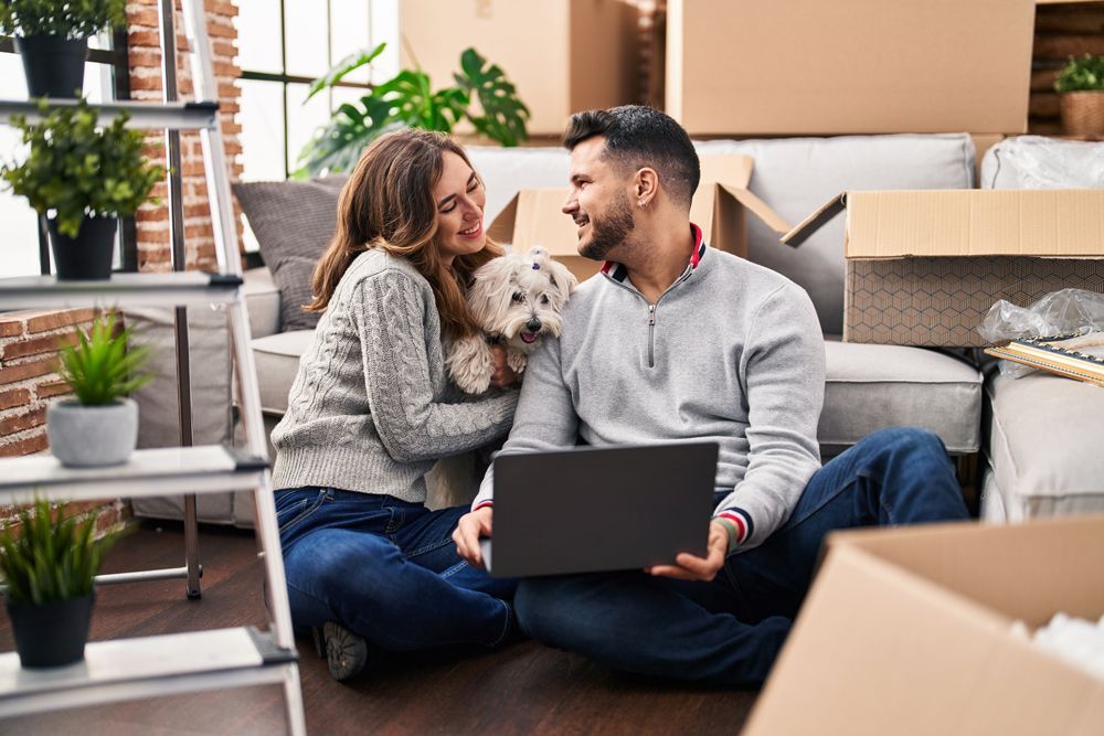 A man and woman are sitting on the floor with a dog and a laptop.