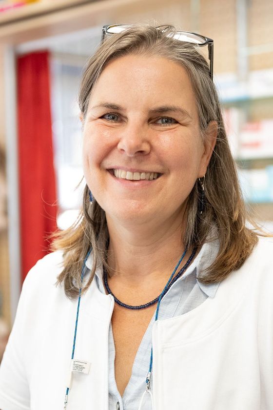 Eine Frau in einem weißen Kittel und mit Brille lächelt in einer Apotheke.
