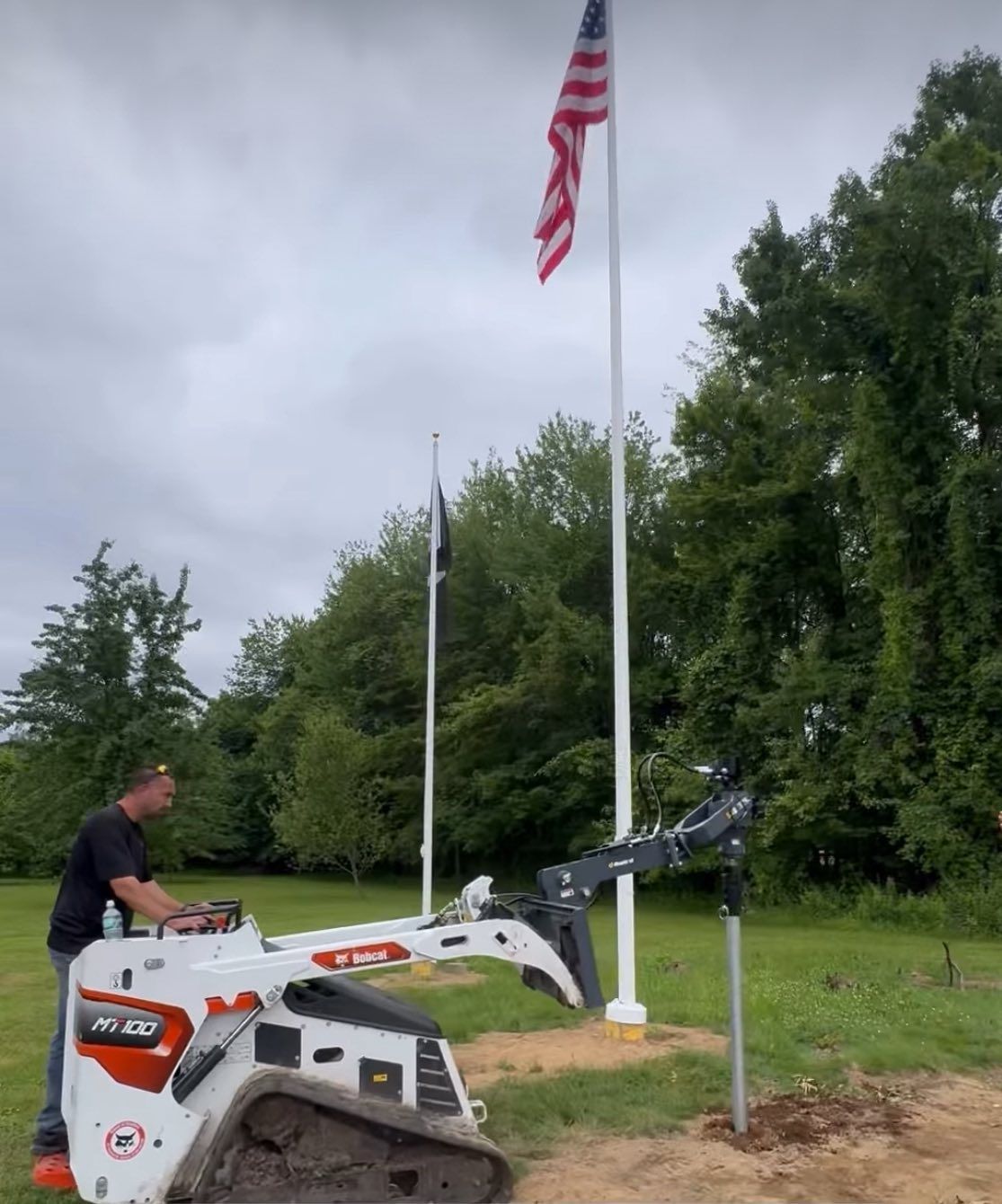Man operating a Bobcat to install a pole. American flag and other flags fly in the background. Outdoors.