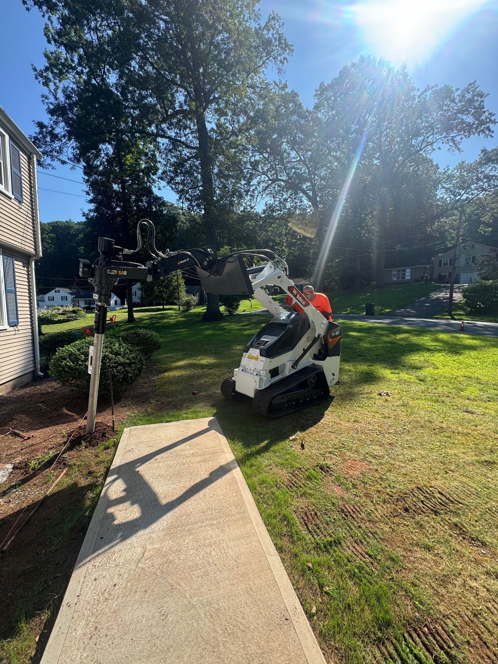 Person operating a white skid-steer on a sunny lawn beside a sidewalk and house