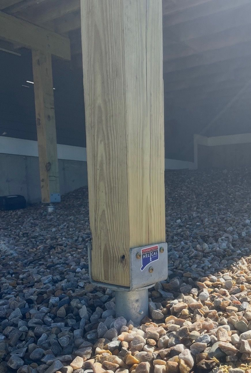 Close-up of a wooden post set in a metal base on gravel beneath an elevated structure.