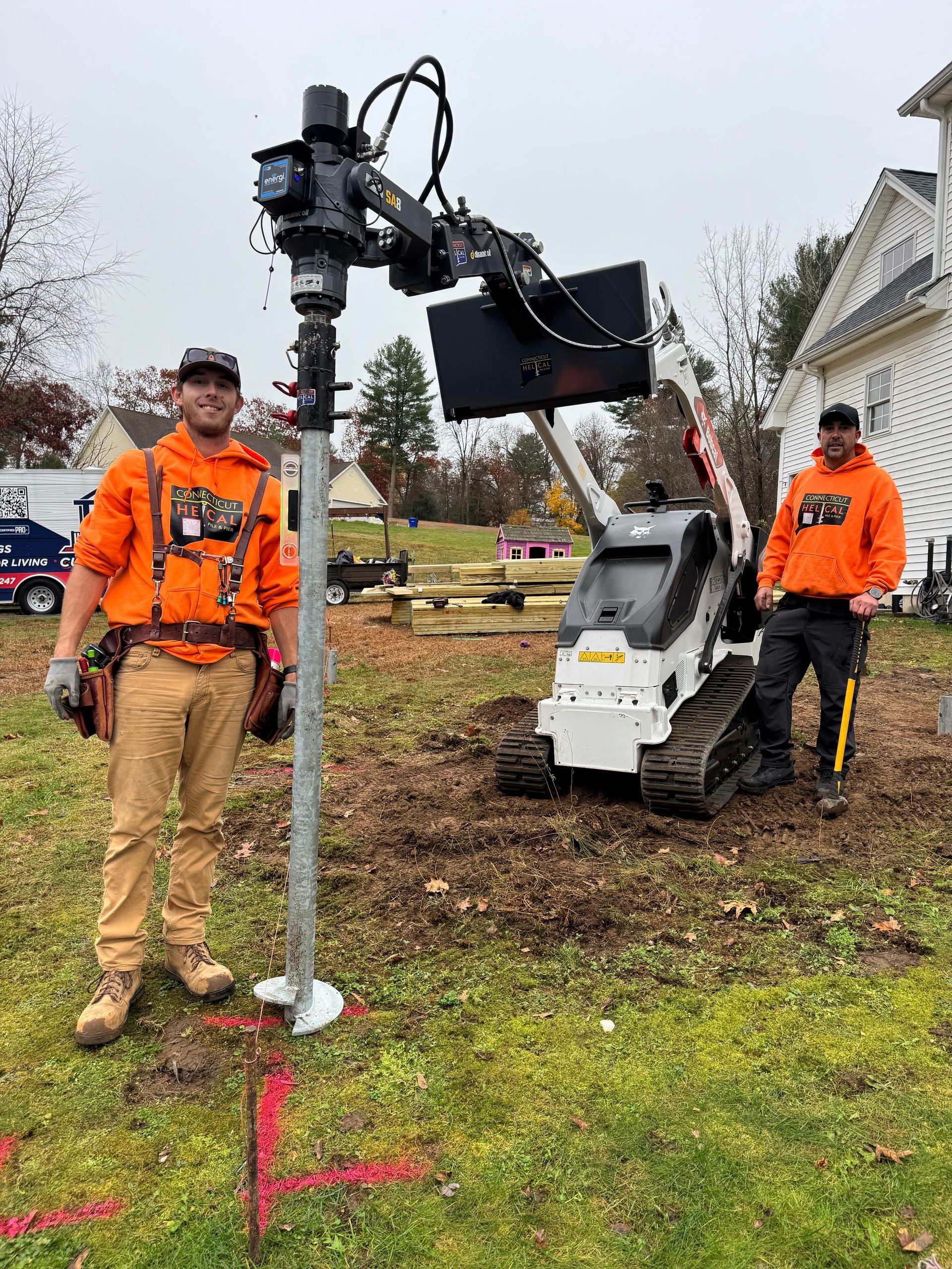 Two workers in orange safety jackets operate a machine-mounted drilling rig outdoors