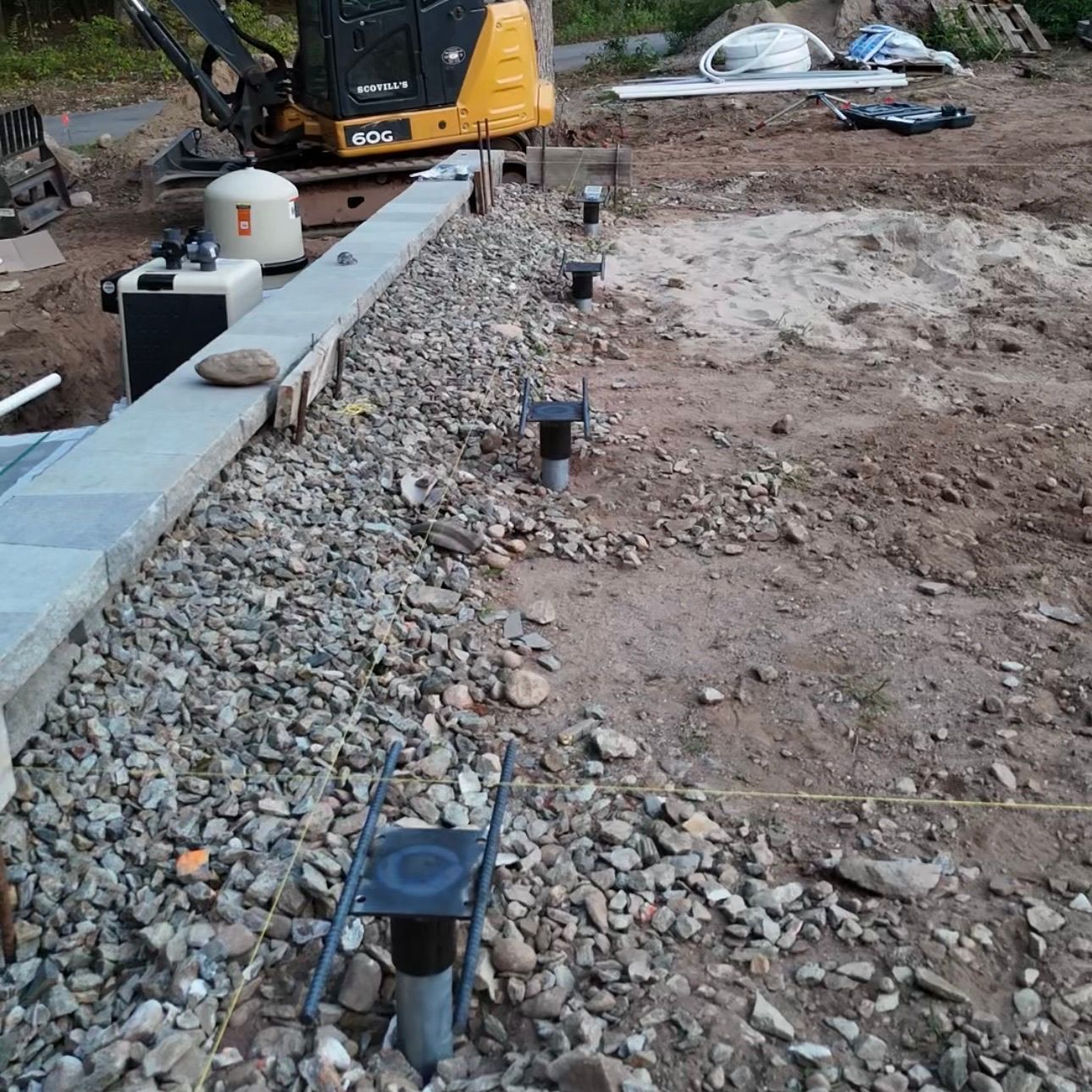 Construction site with gravel trench, black utility posts, and a small excavator in the background.
