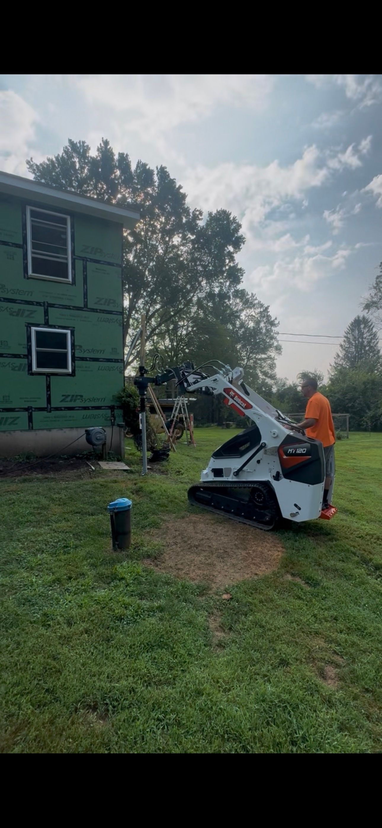 Person operating a white boom lift beside a green house in a grassy yard.