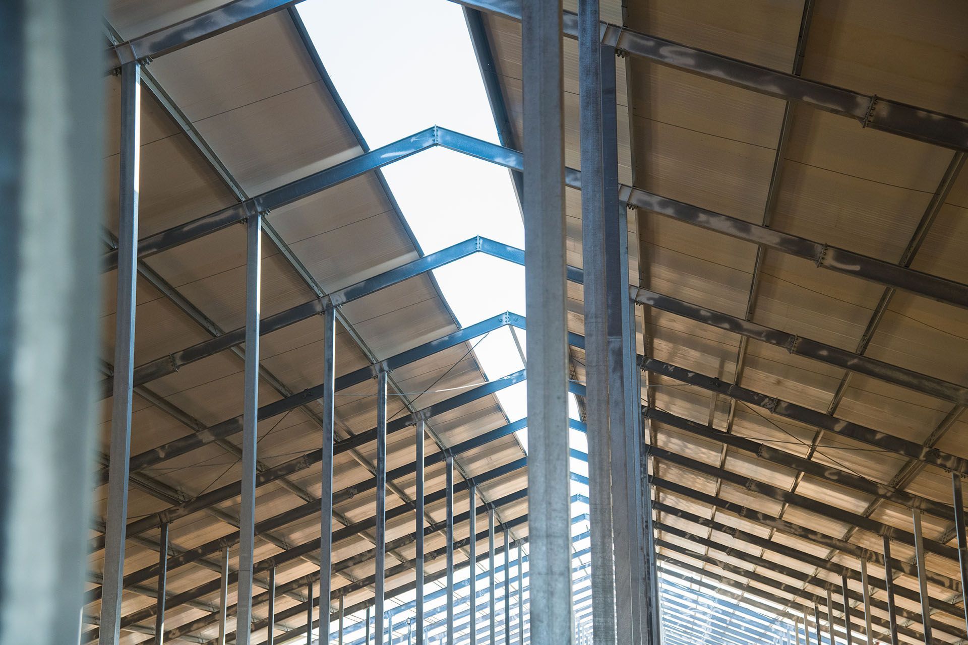 Inside a large warehouse, rows of metal supports hold a corrugated roof with skylights.