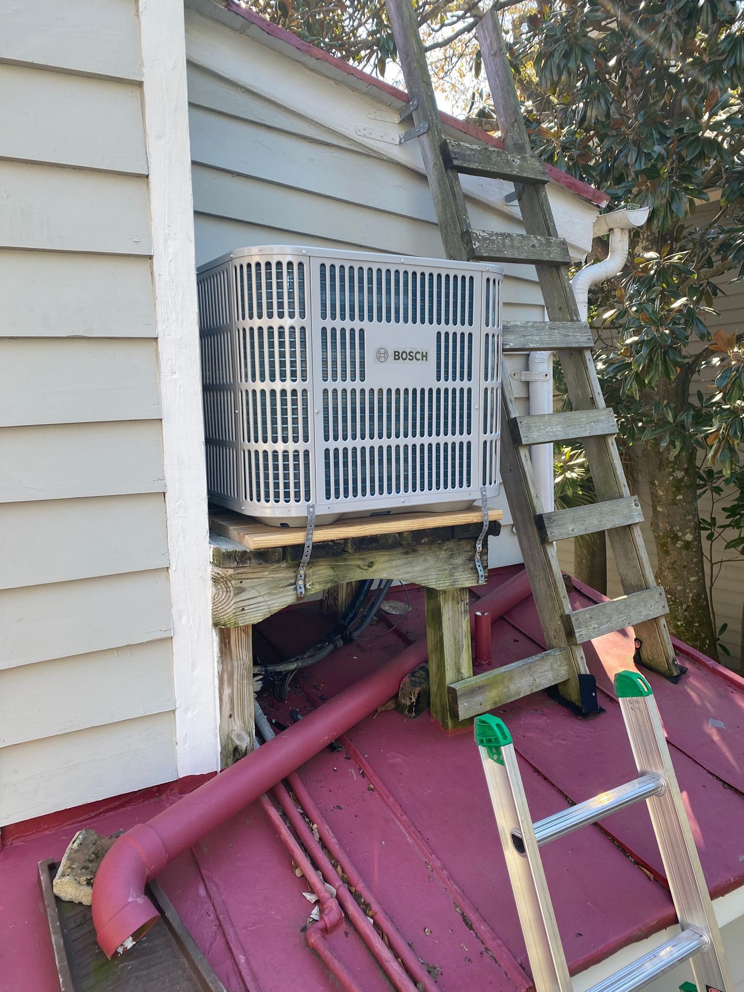 A white air conditioning unit sits on a wooden platform on a red metal roof, next to a wooden ladder leaning on a house.