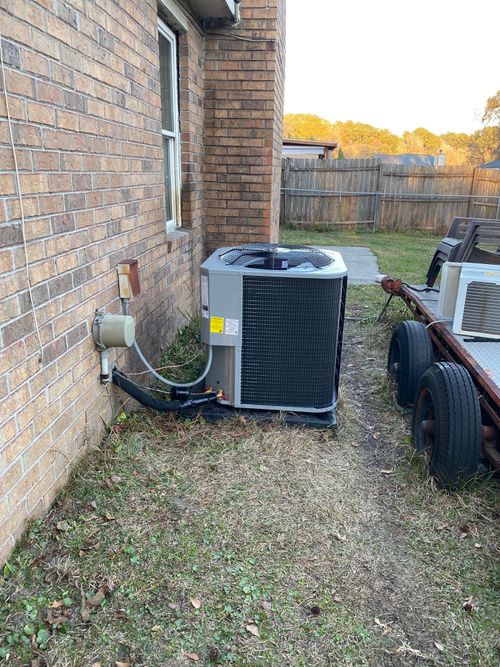 An outdoor HVAC condenser unit sits against a brick house wall near a trailer on a grassy lot.