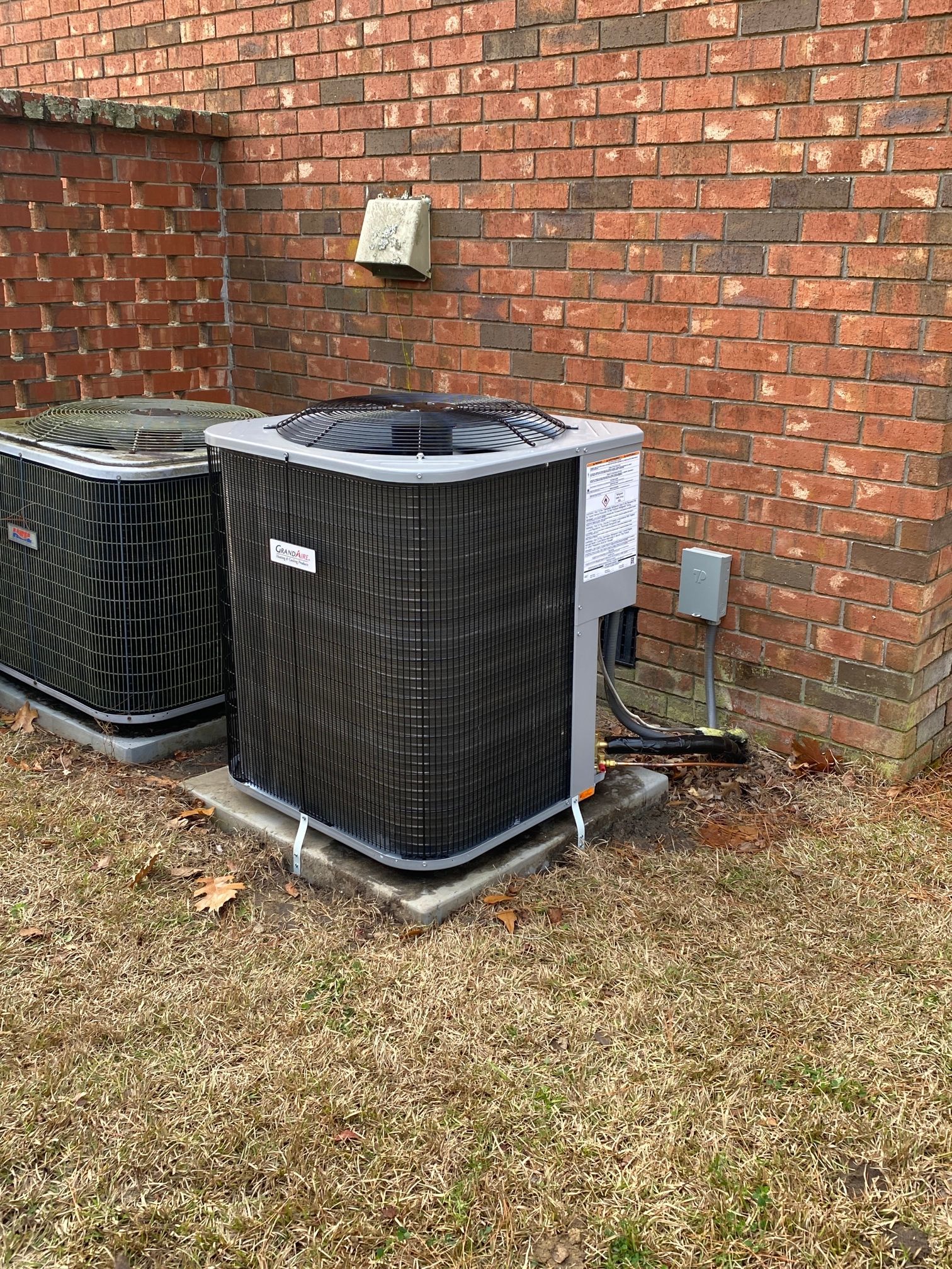 Two gray air conditioning condenser units sit on concrete pads against a brick exterior wall.