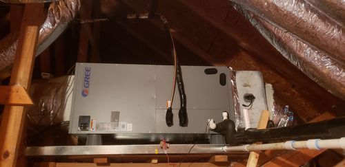 A Gree air handler unit mounted on a metal platform in an attic with insulated ductwork and PVC plumbing pipes.