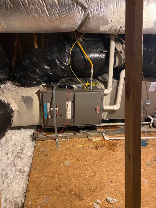 A gray HVAC furnace unit sits in an attic, connected to white pipes and large, black air ducts amidst insulation.