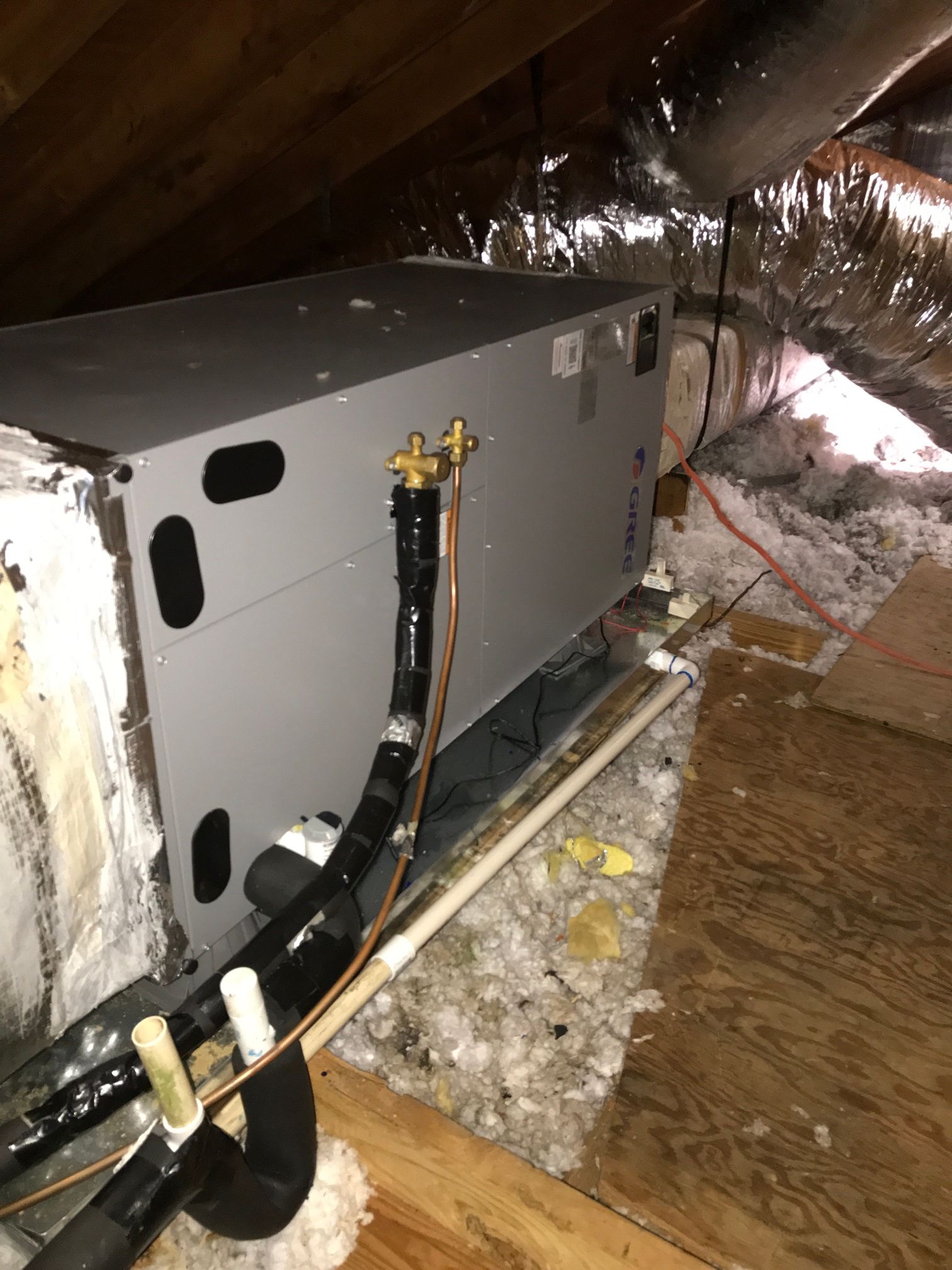 An HVAC air handler unit in an attic, with visible refrigerant lines and piping resting on wooden floorboards.