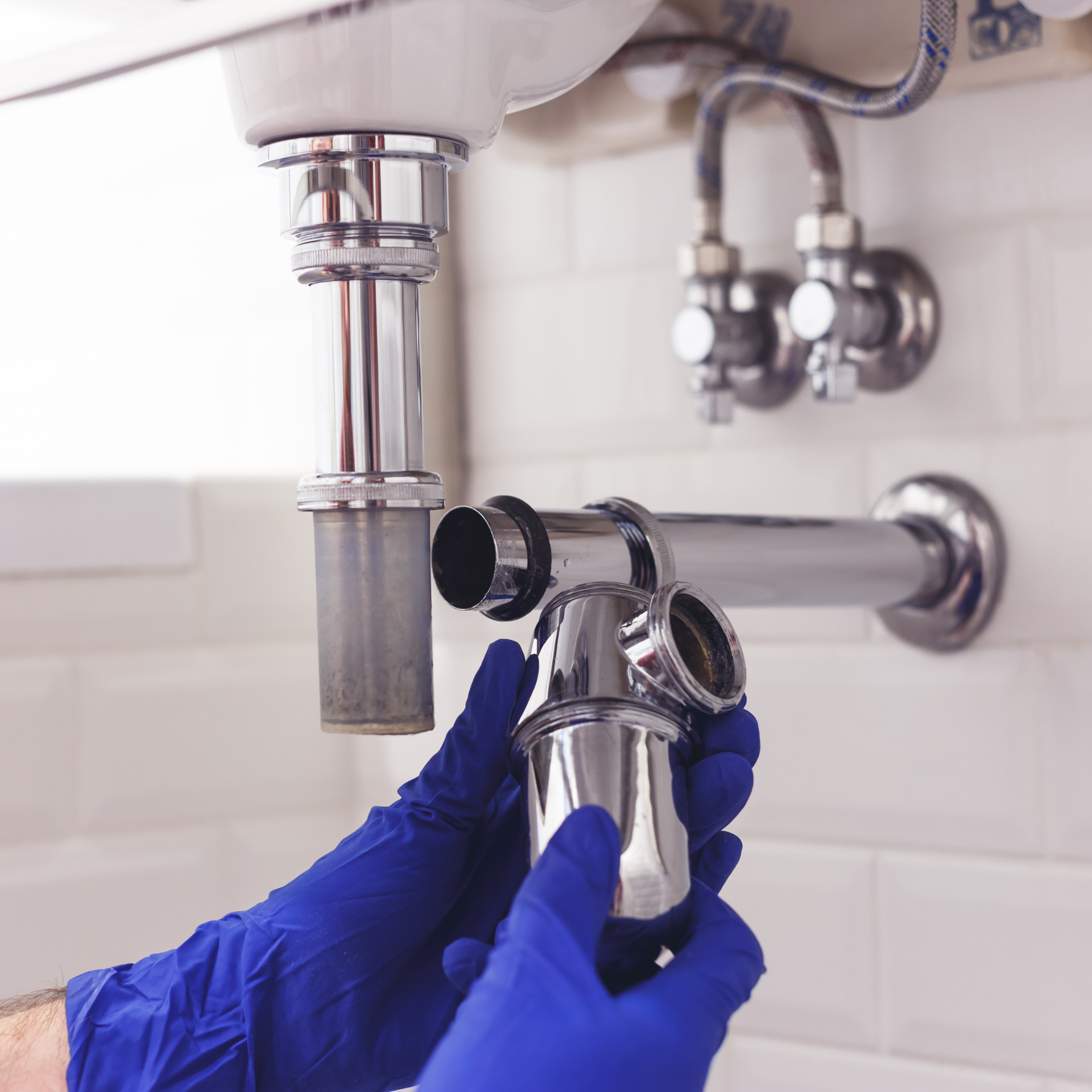 A plumber wearing blue gloves installs a chrome P-trap under a bathroom sink.