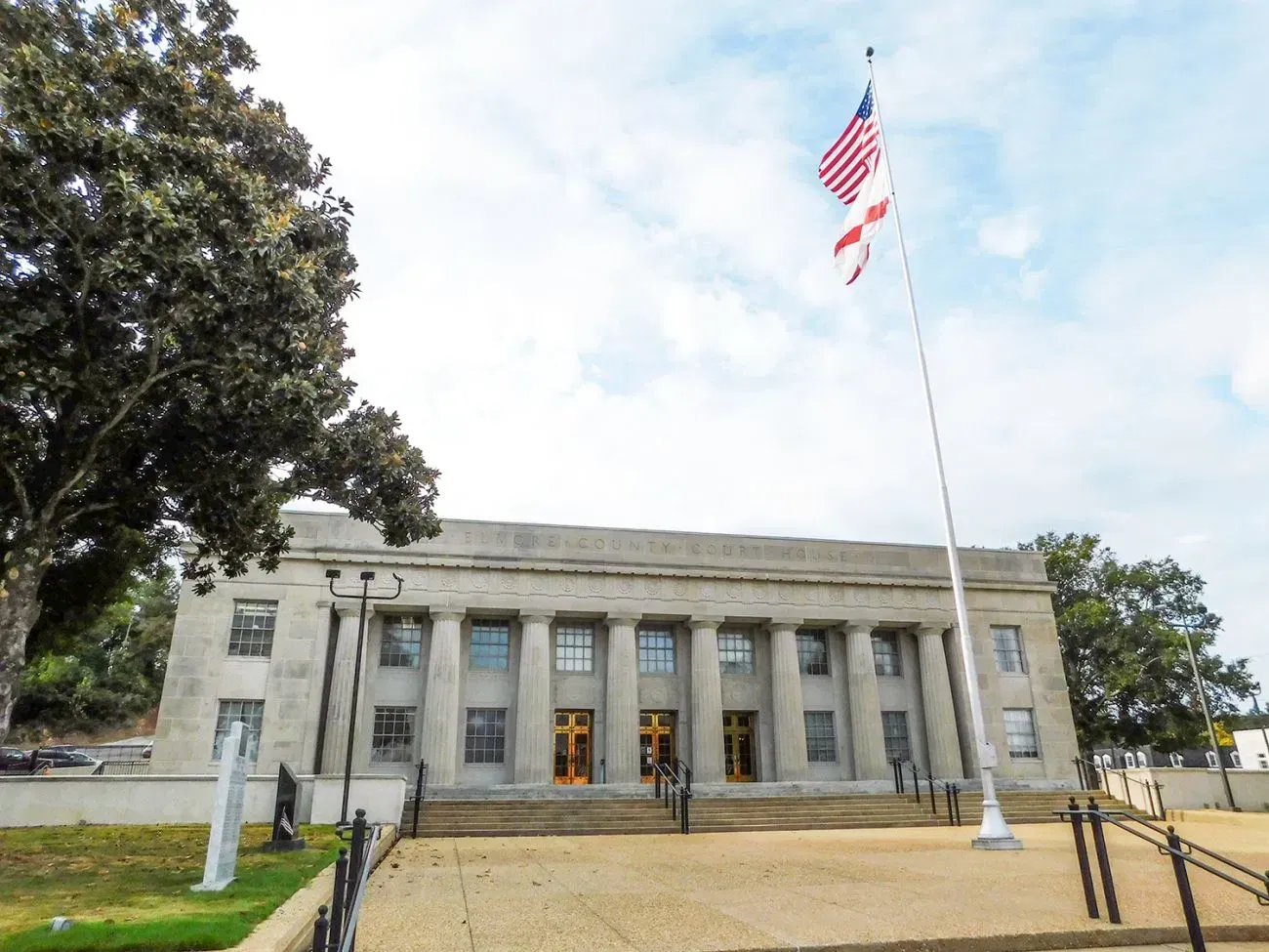 Courthouse with flag at half-mast; light-colored stone building with columns, under cloudy sky.