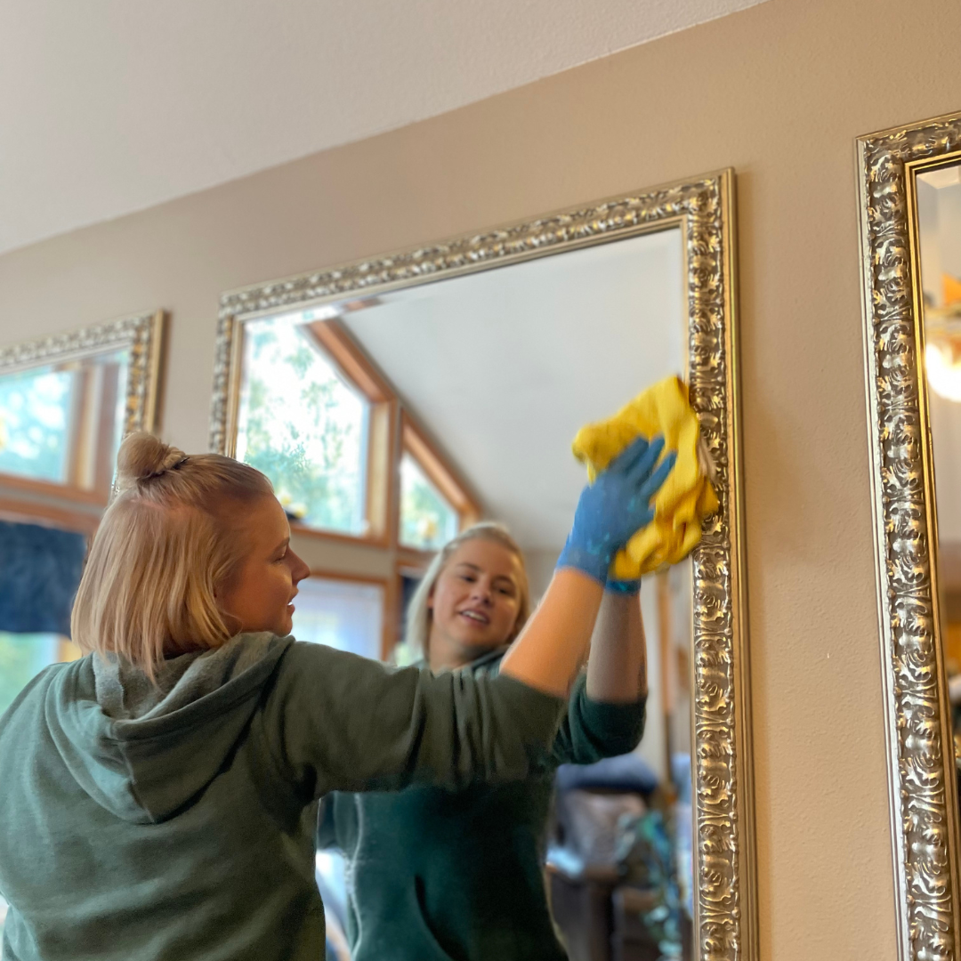 Woman cleaning a large mirror with yellow cloth, wearing a green hoodie and blue gloves, indoors.