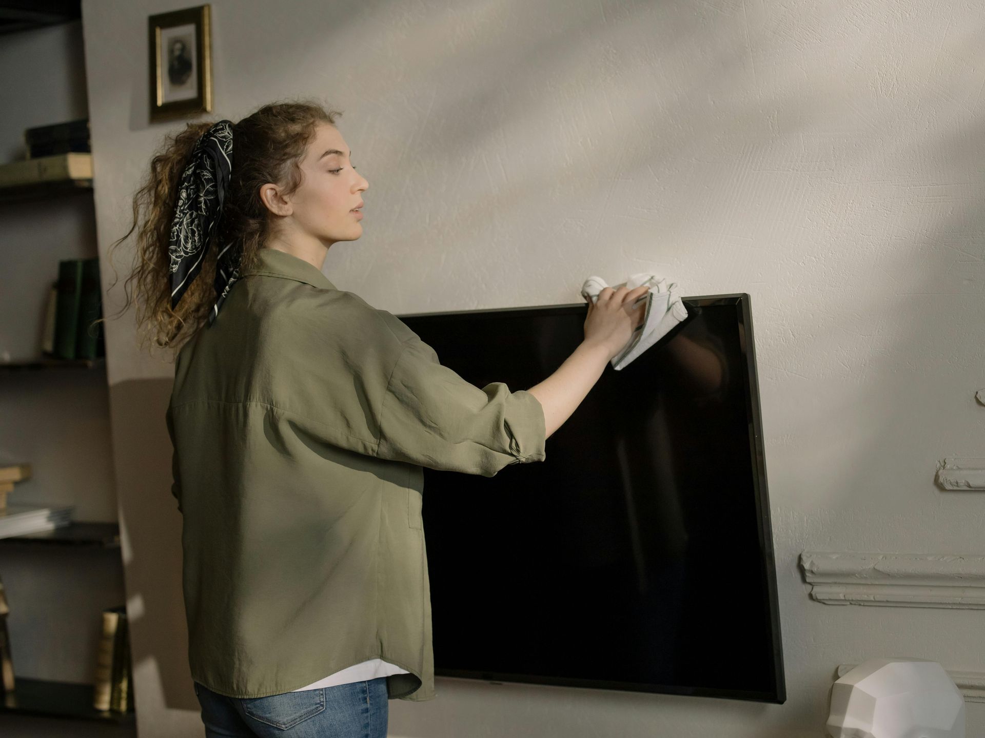 Woman cleaning a mounted television screen with a cloth in a living room.
