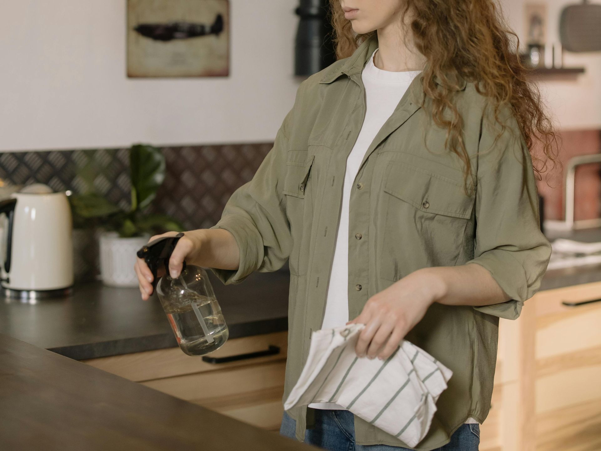 Woman spraying a countertop with a spray bottle, holding a cloth in a kitchen.