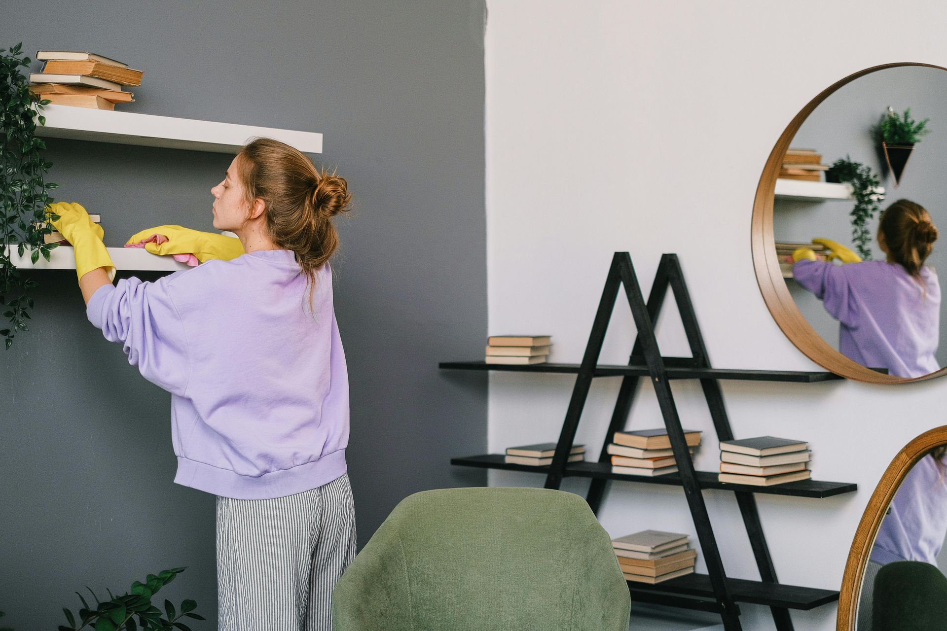 Woman wearing yellow gloves cleaning shelves with plants and books.
