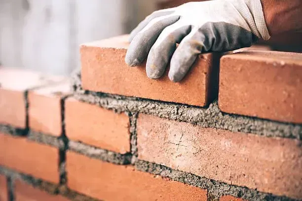 Gloved hands placing red bricks on a brick wall with mortar