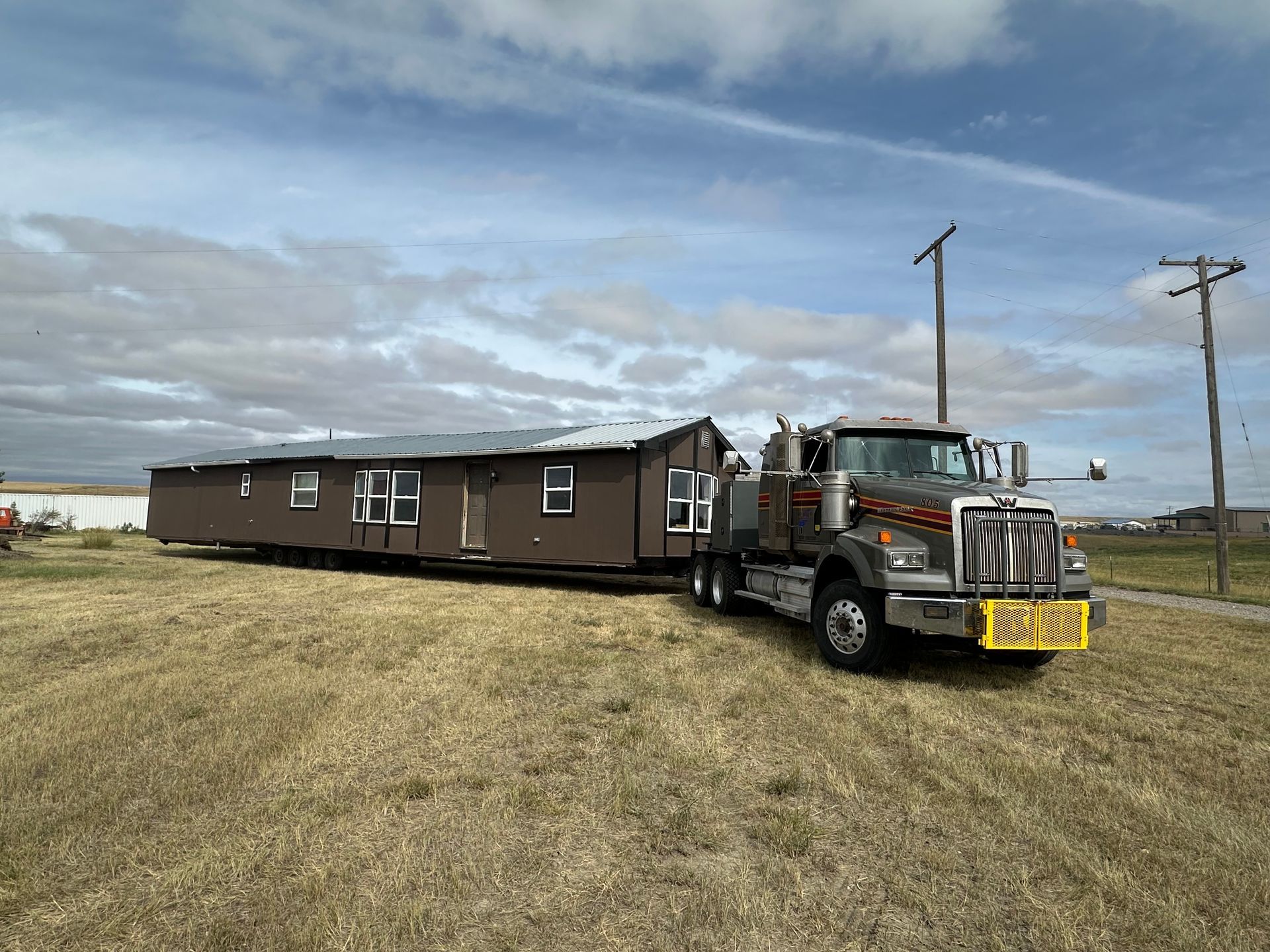 Long brown mobile home being towed by a semi-truck across a grassy field under a cloudy sky
