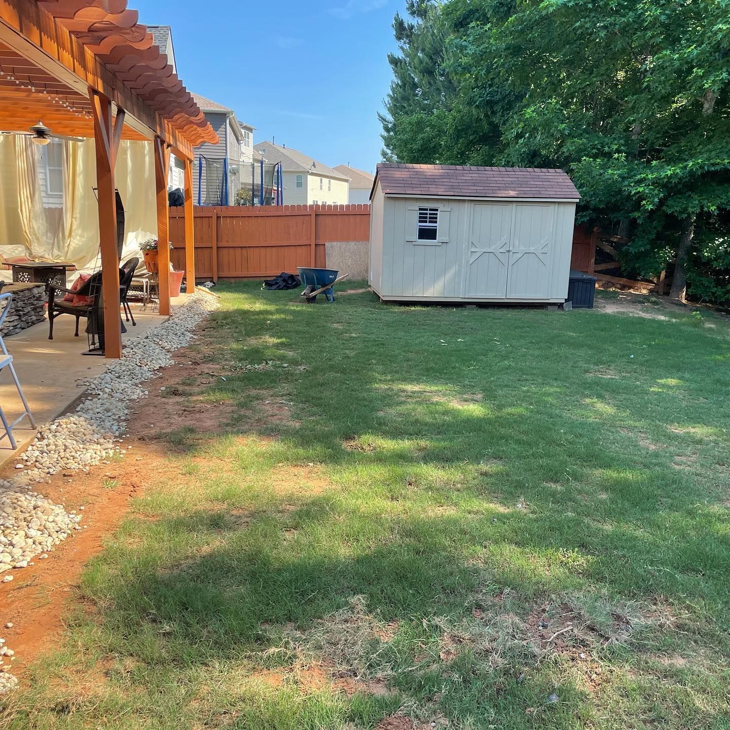 Backyard with a small white shed, green lawn, wooden patio, and large trees on a sunny day