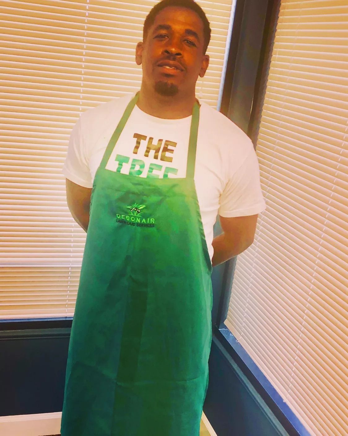 Man standing indoors in a green apron and white T-shirt, posed in front of window blinds.
