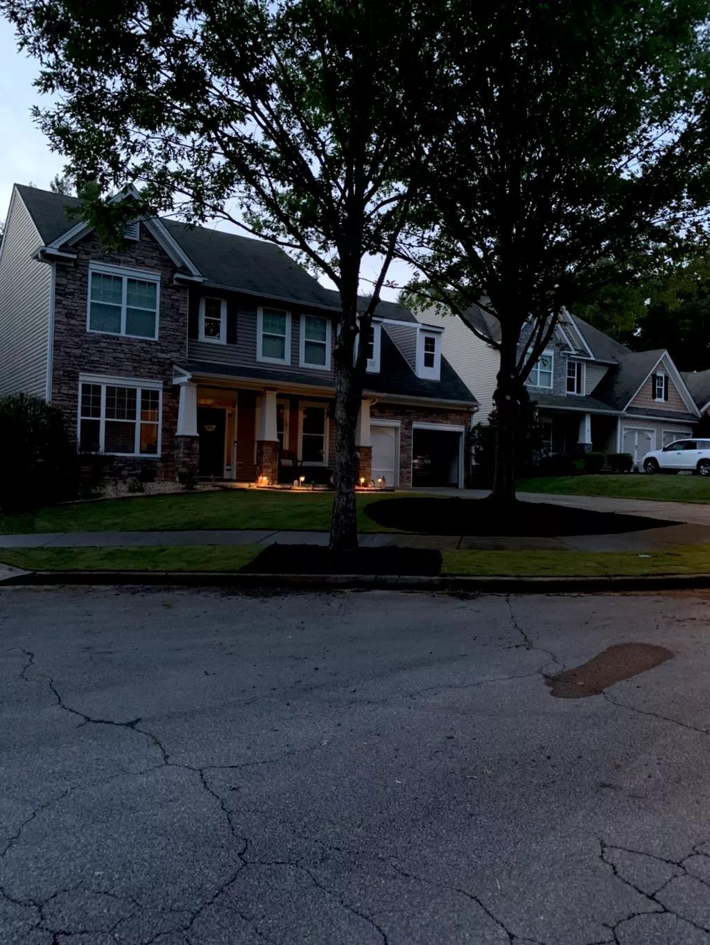 Quiet suburban street with cracked pavement, trees, and lit two-story houses at dusk.