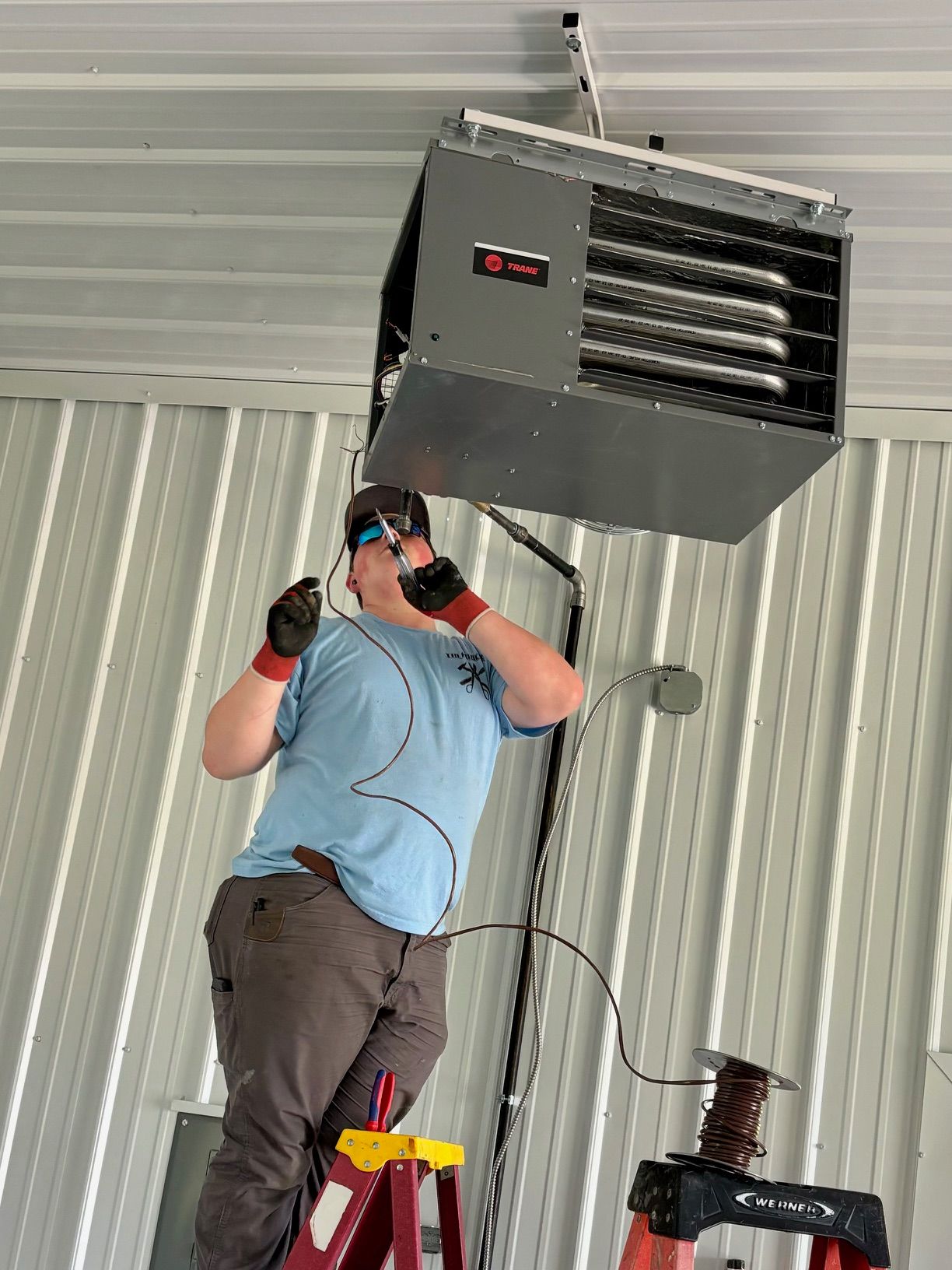A man is standing on a ladder working on a heater hanging from the ceiling.