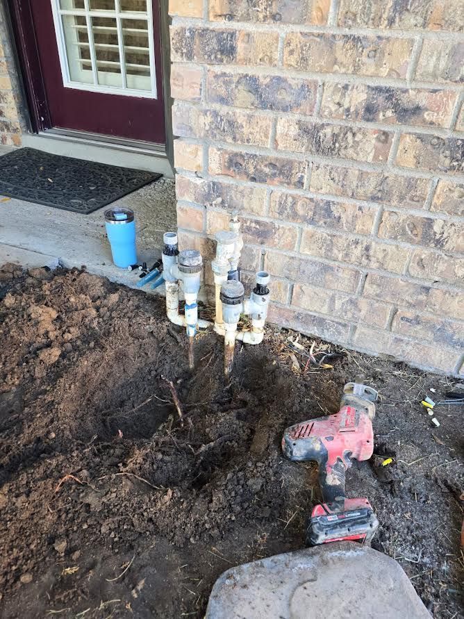 Exposed utility pipes beside a brick house, with a power drill on the dirt near the foundation.