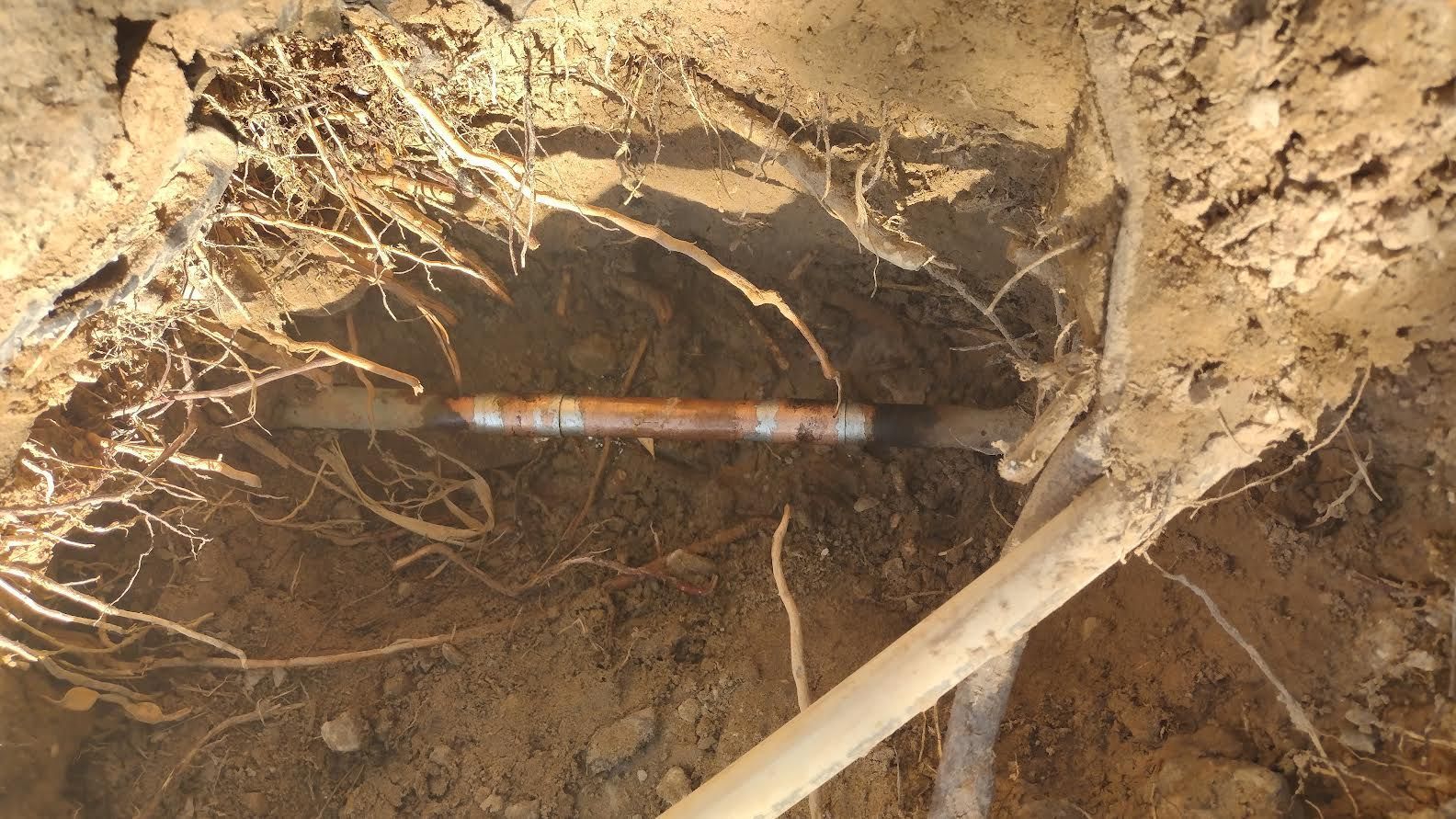 Partially buried utility pipe in a dirt trench with exposed roots and a shovel handle nearby