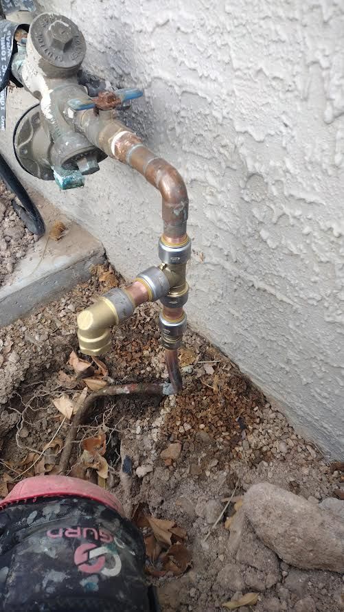 Outdoor copper water pipes and valves against a stucco wall, with debris on the ground nearby