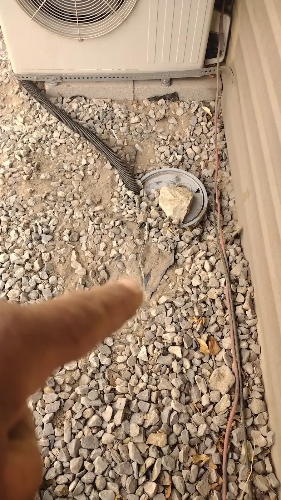 Hand pointing at a damaged outdoor cable and round fixture on a rocky ground beside a building wall