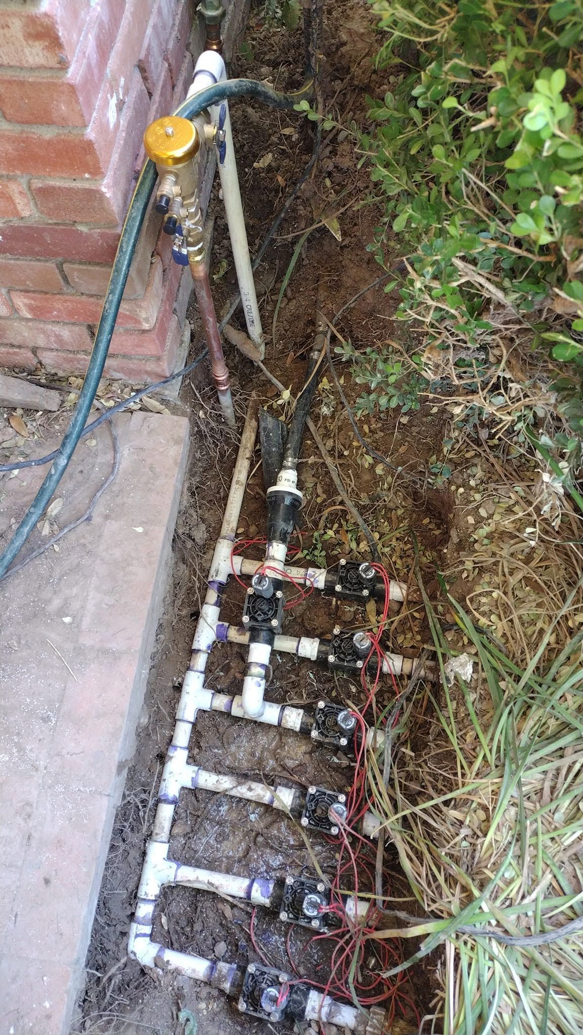 Outdoor utility valves and pipes beside a brick wall, with water meter boxes and overgrown plants.