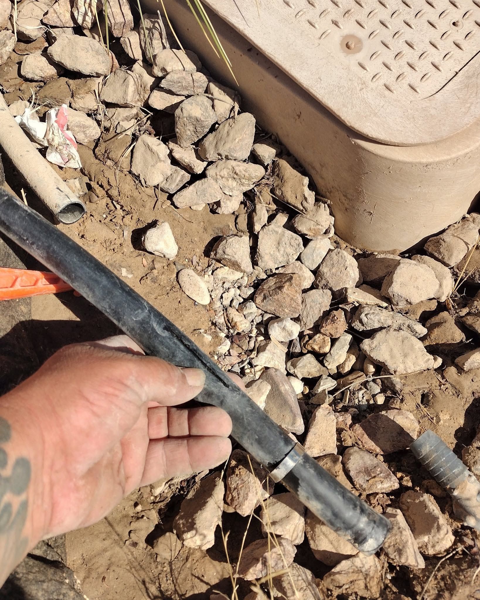 Hand holding a black pipe over gravel beside a tan vent cover