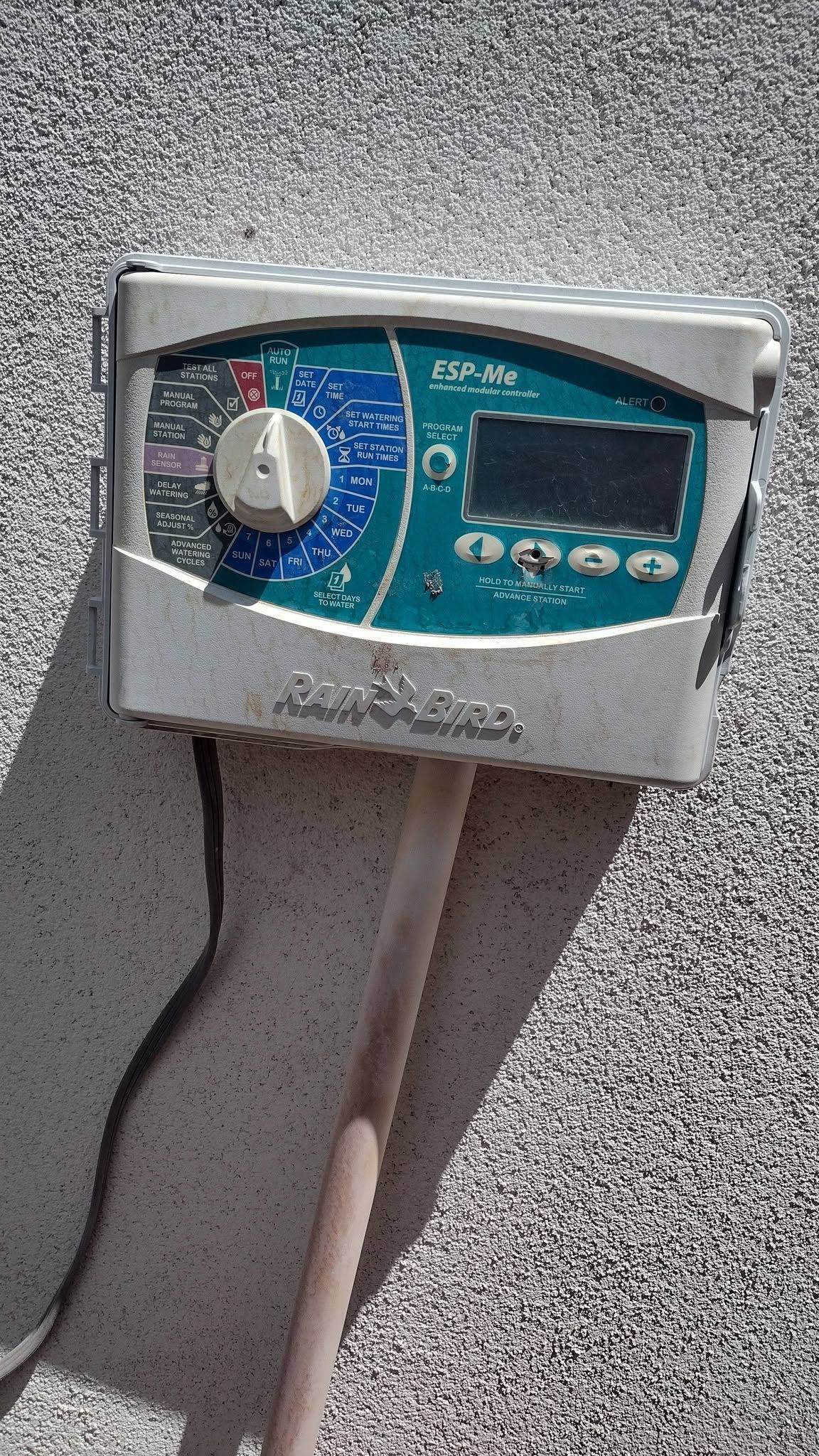 Wall-mounted digital timer with a knob and display, attached to a stucco exterior wall with cables underneath