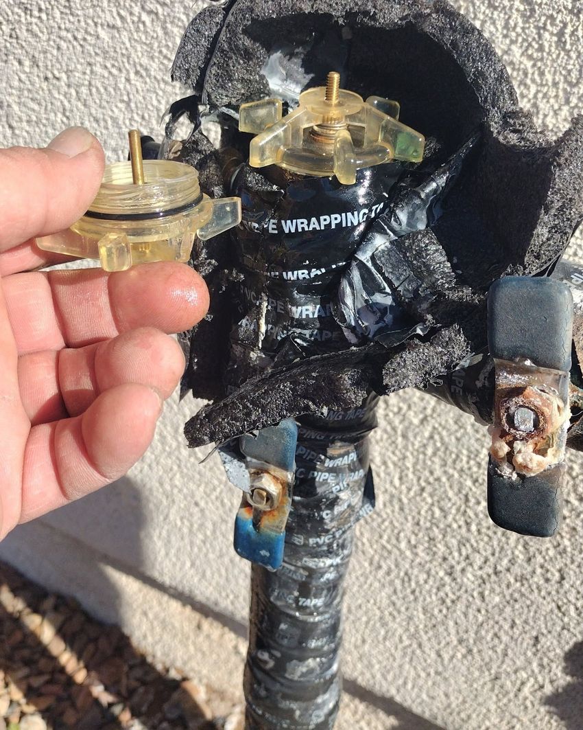 Hand holding a broken brass faucet valve beside exposed outdoor plumbing wrapped in black tape