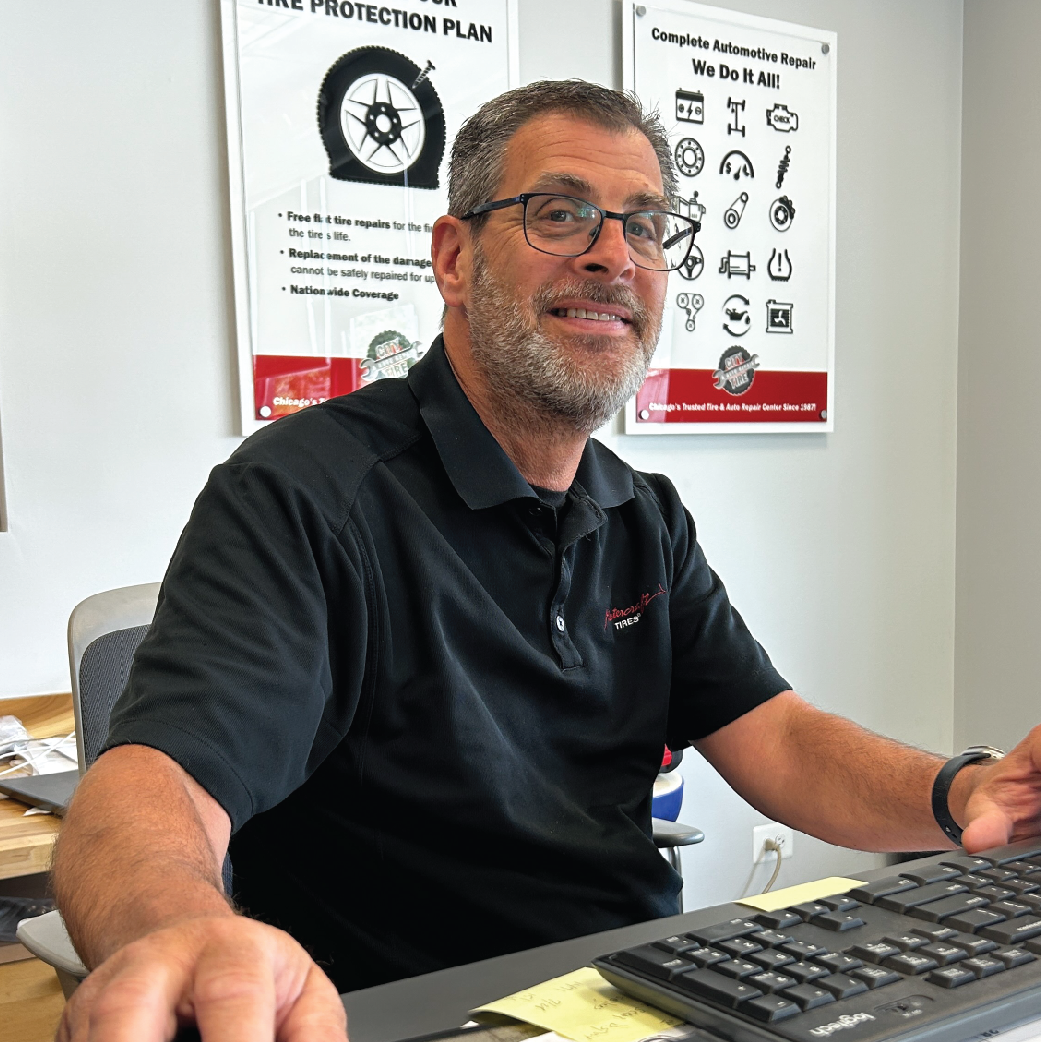 Man with glasses, black shirt, seated at a desk in an office setting with posters on the wall.
