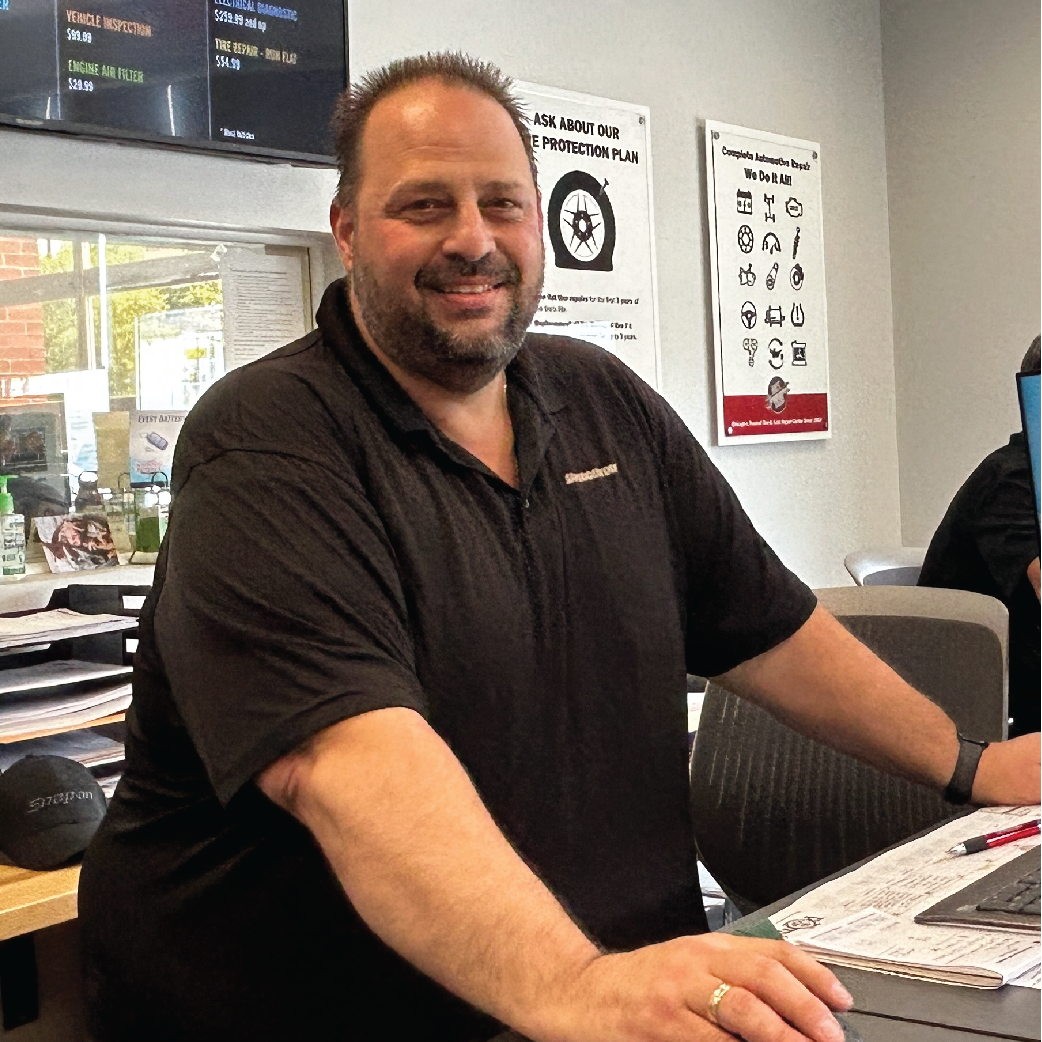 Man with beard smiles at a desk, wearing a black shirt. Office setting.