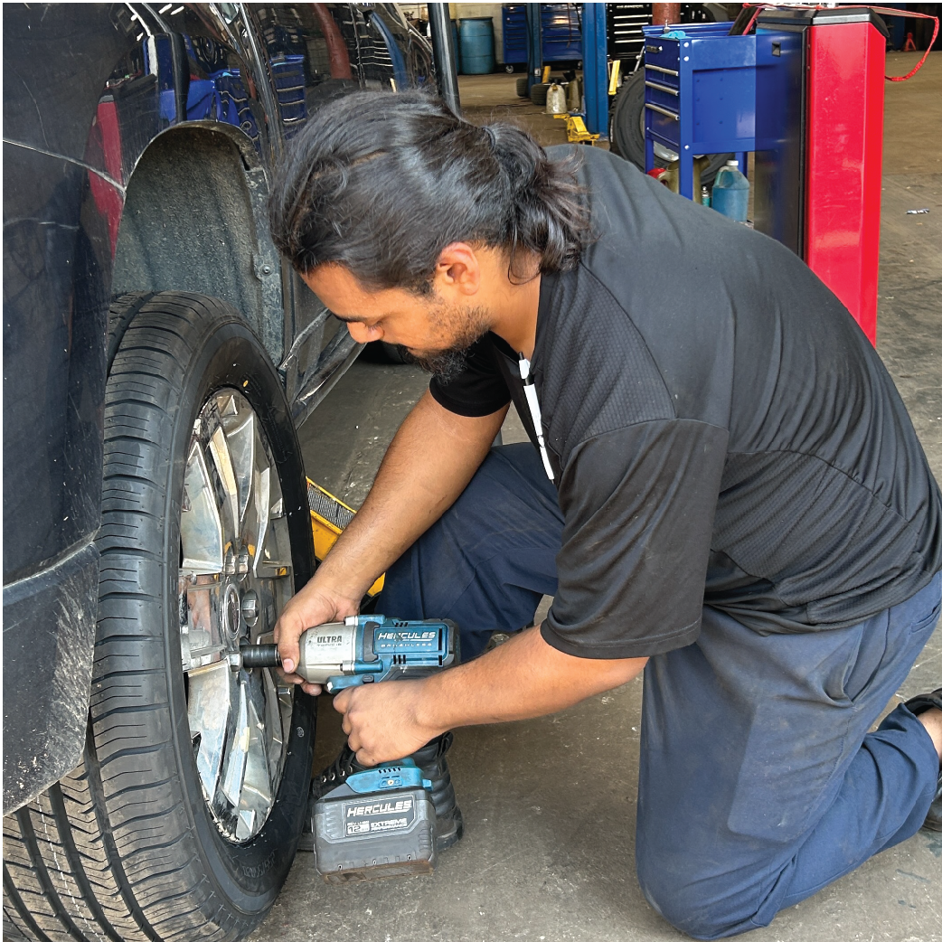 Mechanic kneeling, using a power tool to remove a tire from a car. Garage setting, blue and black colors.