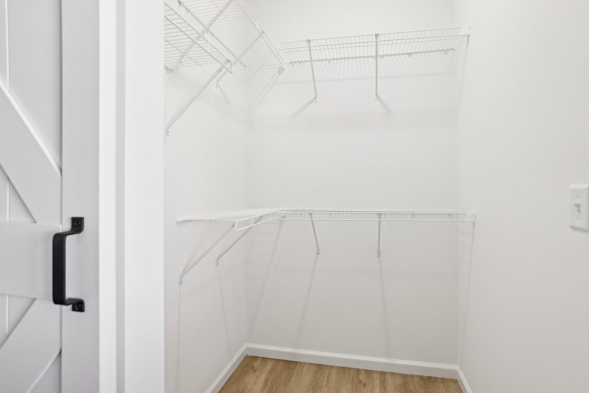 Empty white walk-in closet with wire shelves and hanging rods, wood-look floor, and black door handle