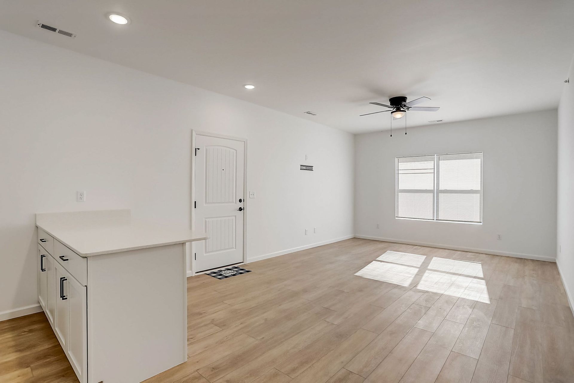 Bright empty living room with white walls, light wood floors, and sunlight through a window blinds.