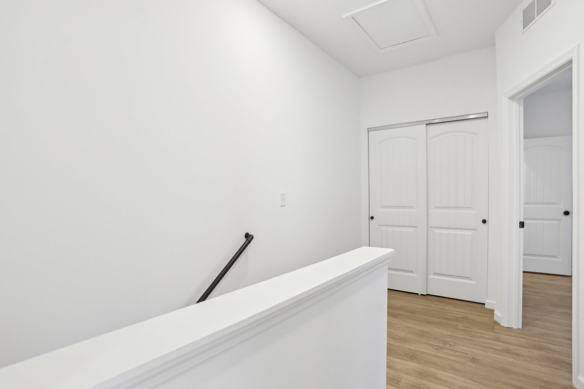 Bright white hallway with wood flooring, stair railing, and closed double doors at the end