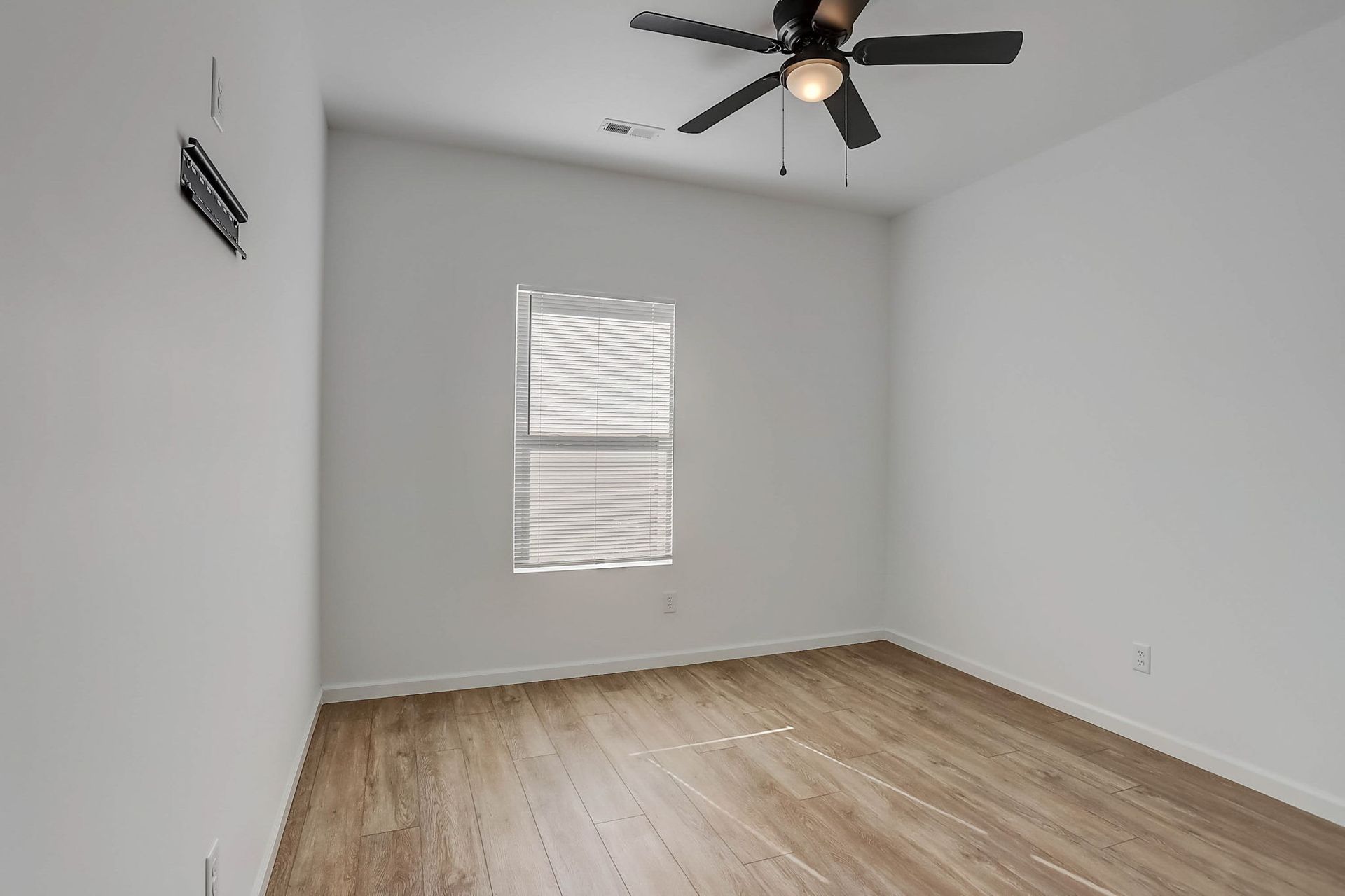 Empty white bedroom with wood floors, a ceiling fan, and a window with blinds.