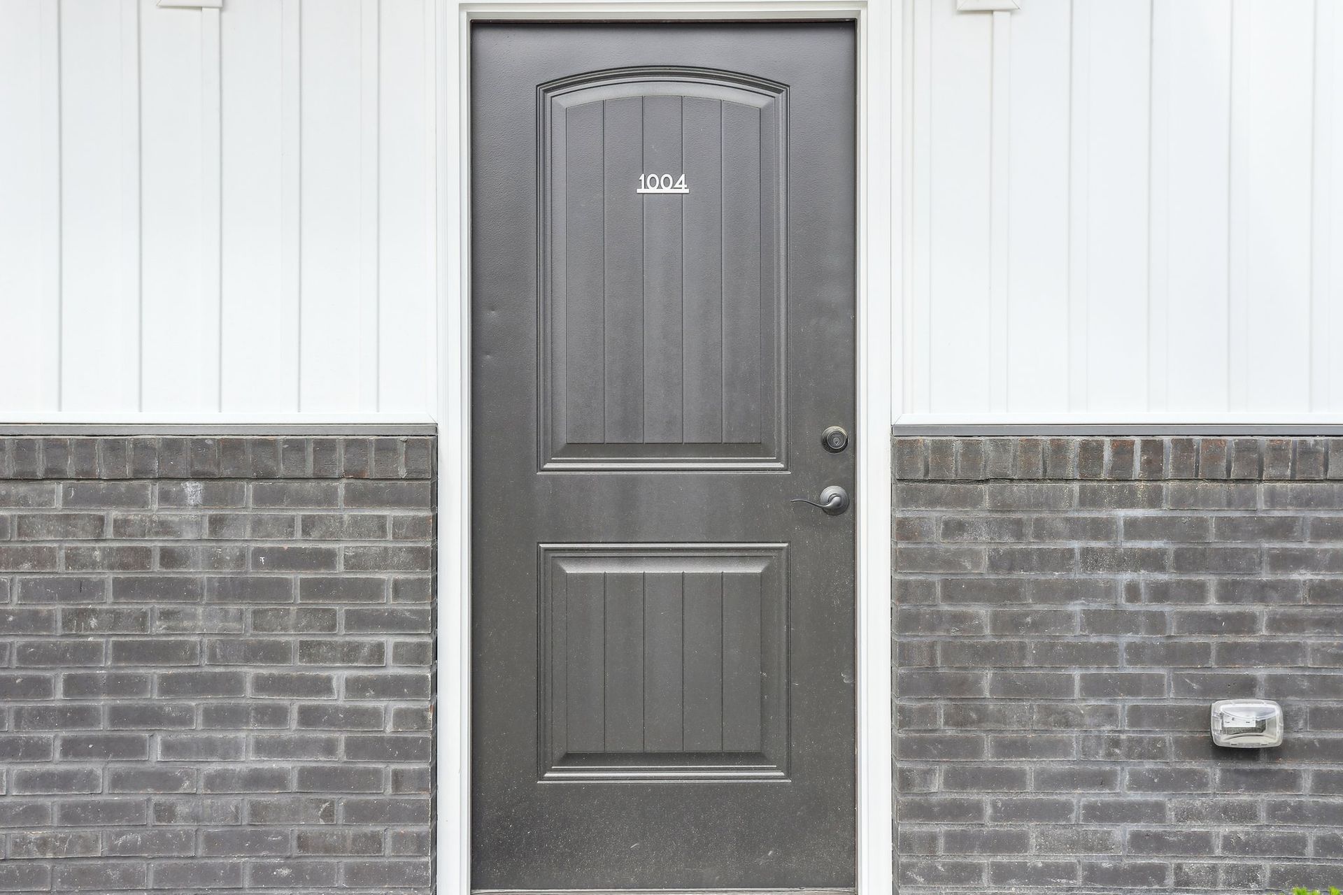 Gray front door with a small number plaque, set in a white and brick exterior wall.